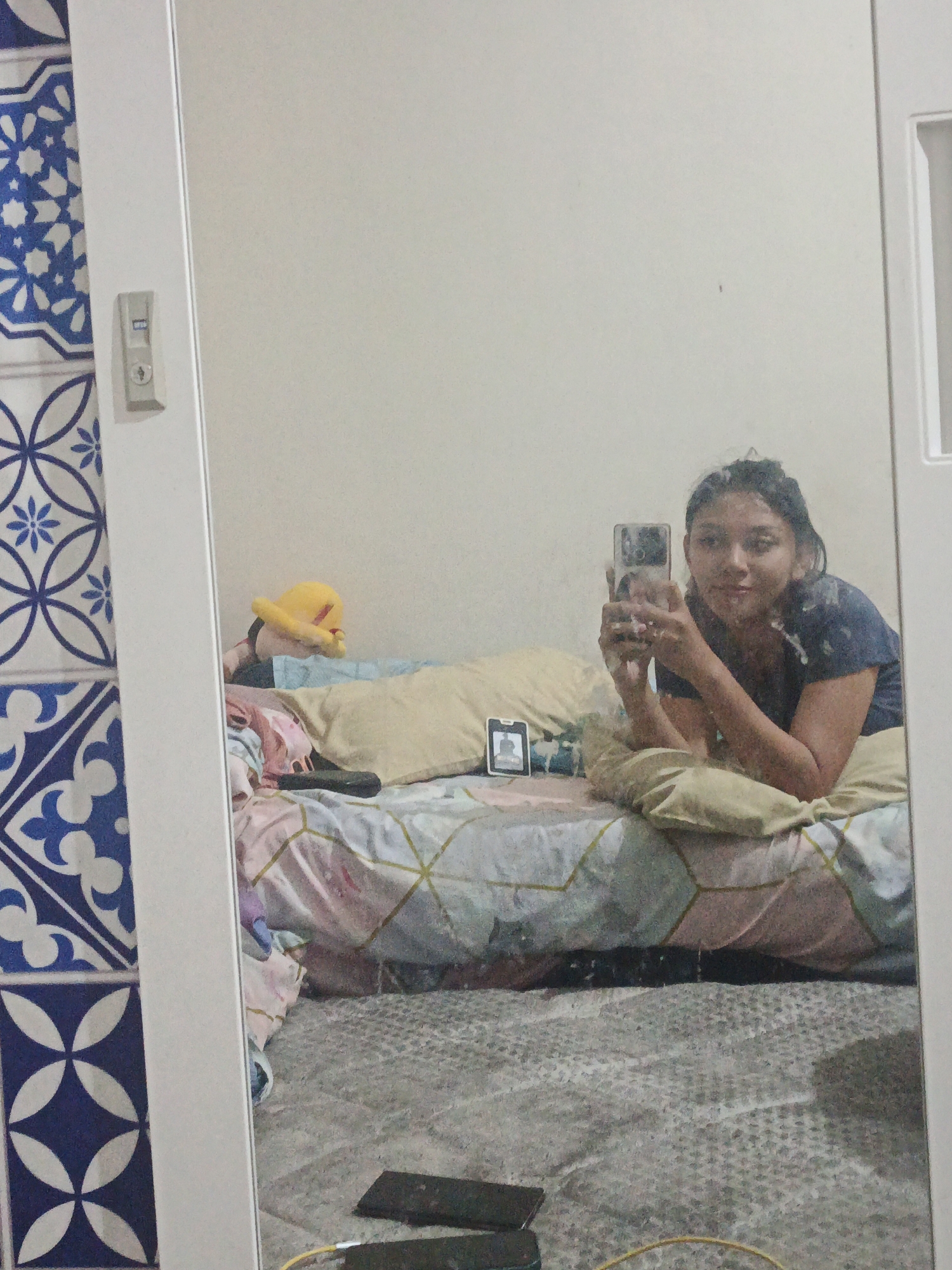 A young woman is taking a mirror selfie in what appears to be a bedroom. She is lying on a bed with patterned sheets and multiple pillows. Behind her, a stuffed animal with a yellow head and red face is visible. To the left of the mirror, a section of a wall with blue and white patterned tiles is shown, and a light switch is mounted on the white frame of the mirror. The woman is wearing a dark blue t-shirt and is holding a smartphone in her right hand to capture the reflection. The overall environment suggests a casual, domestic setting, possibly a dorm room or apartment. The image is taken from a slightly high angle, looking down at the woman and the bed. The lighting is soft, indicating indoor illumination.