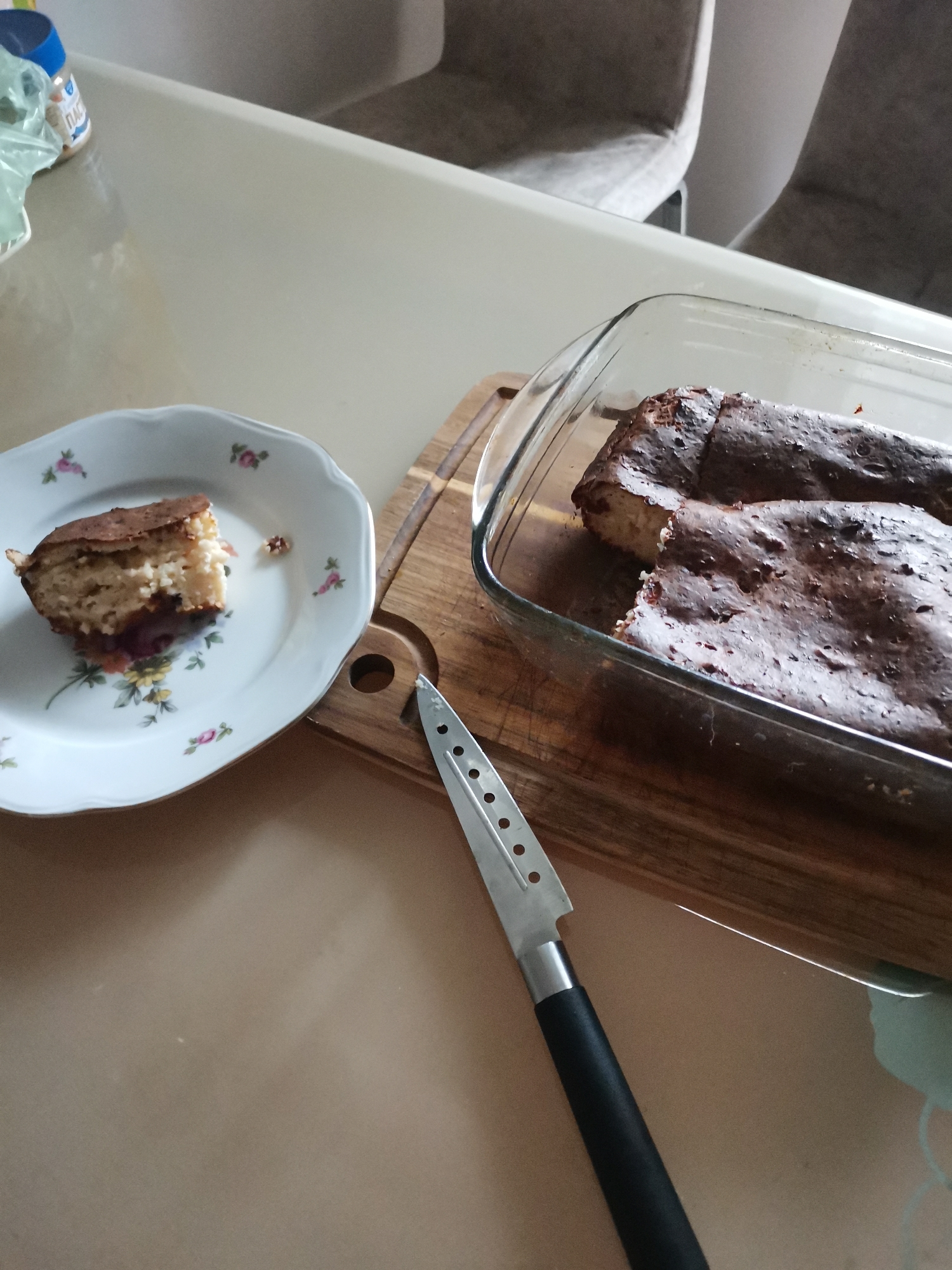A slice of cake with berry filling sits on a white floral-patterned plate next to a glass baking dish containing a partially eaten cake. A silver knife with a black handle rests on a wooden cutting board between the plate and the baking dish. The scene is set on a light-colored table, with a blurred background of chairs. The location is a domestic kitchen in Chaikovsky, Russia.