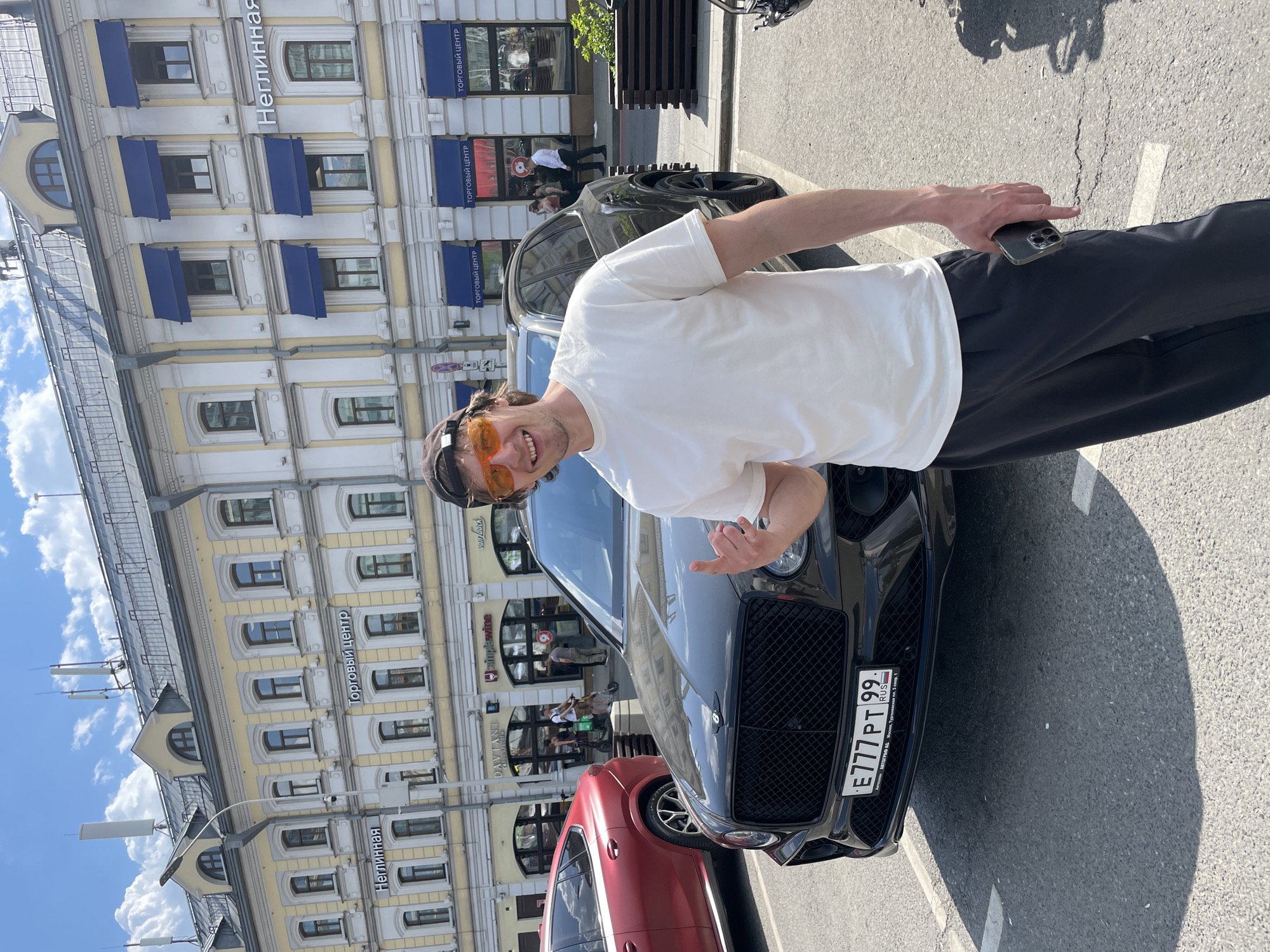 The image shows a man in a white t-shirt, black pants, and orange sunglasses standing next to a black Bentley. He's holding a phone in one hand and making a hand gesture with the other. A red car is partially visible next to the Bentley.
The location appears to be in Moscow, Russia, based on the license plate of the Bentley, which reads "E777 PT 99 RUS." The building in the background has awnings and signs in Russian, including "Неглинная" and "Торговый Центр" (Shopping Center), suggesting that the location is Neglinnaya Street in Moscow. Businesses are also visible behind the man. The photo appears to be taken on a sunny day.