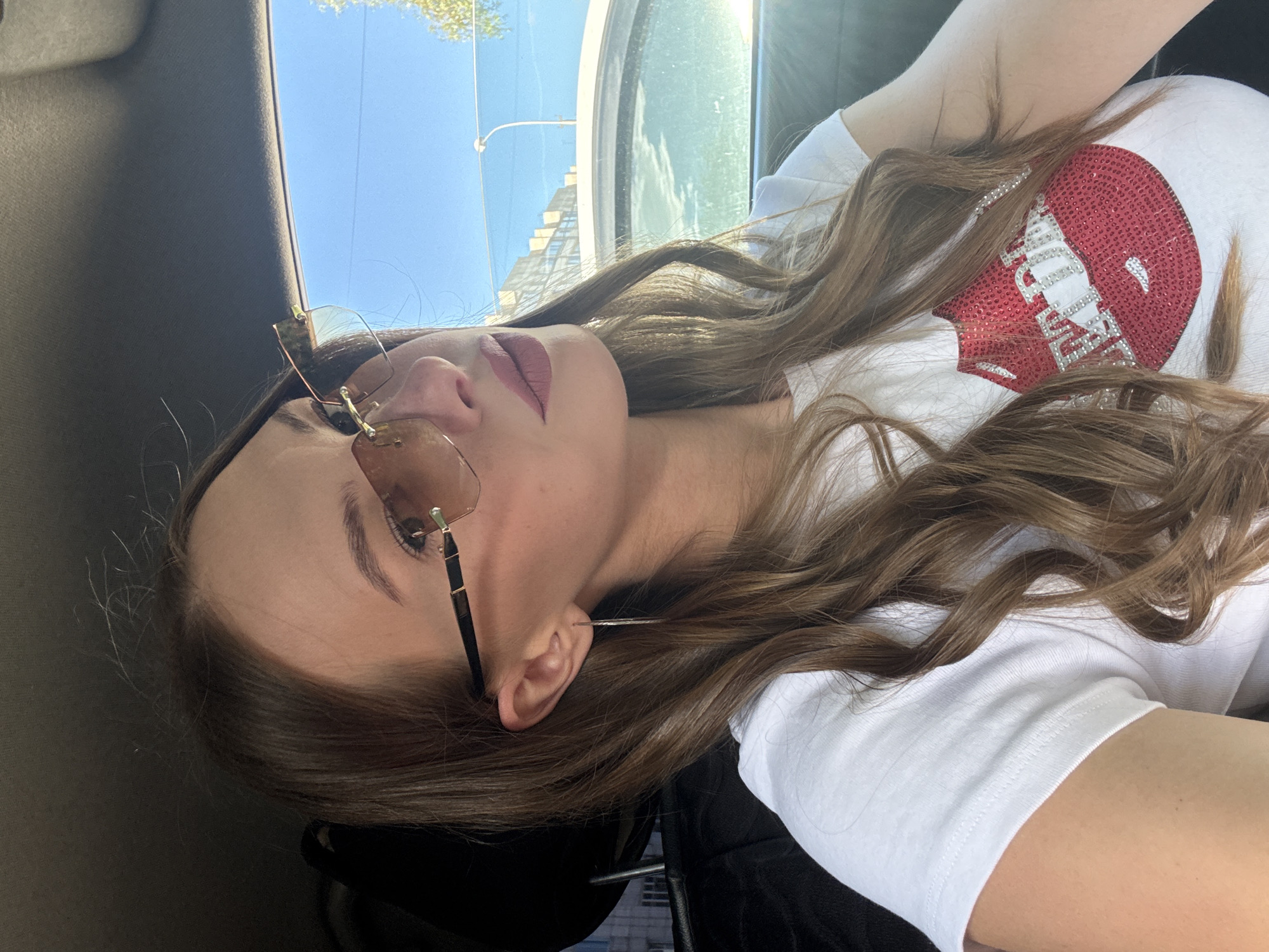 The image shows a young woman wearing sunglasses and a white t-shirt with a red sequined lip design. She is sitting in the passenger seat of a car, with her hair down and parted to the side. The background shows a blurred view of buildings and a clear blue sky, suggesting it is daytime. The setting appears to be a city, likely on a sunny day. There are no specific landmarks visible that would definitively place the location in Perm, Russia.