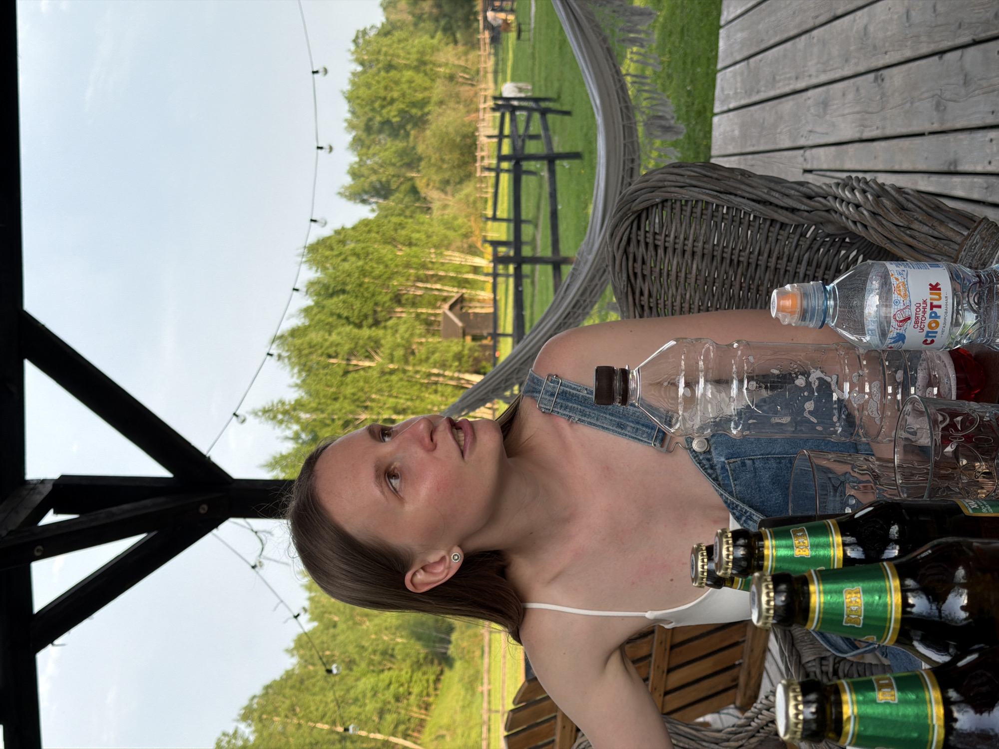 A woman is seated at a table outdoors, likely in Prudki, Russia, with bottles of beer and water. She is wearing denim overalls over a white top. A table made of wooden slats is adjacent to a wicker chair. The background shows a green landscape with trees and a fence.