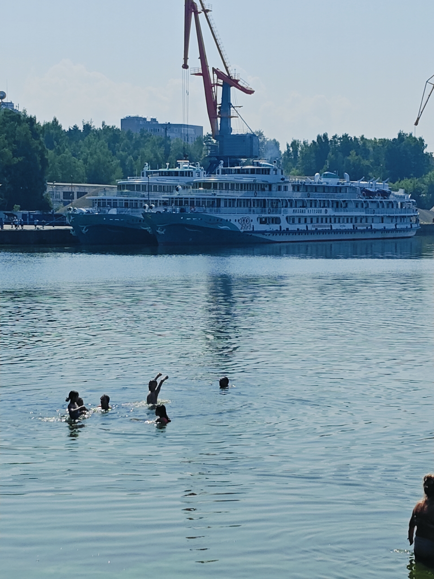 A group of people are swimming in the Kama River in the city of Chaikovsky, Russia, on a sunny day. In the background, two large passenger ships are docked at a port, with a red crane visible above them. The water is calm, with small ripples. The people in the water are of various ages and are engaged in swimming and playing. One person is standing in the water on the right side of the frame, with their back to the camera. The sky is clear and blue, with a few white clouds. The scene suggests a leisurely summer day at the river.