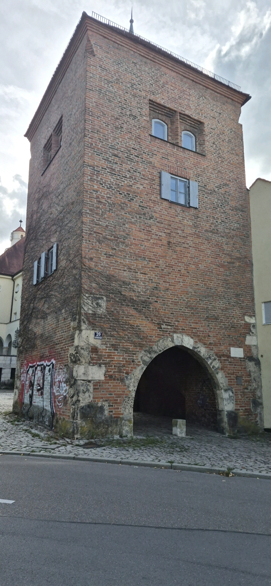 The image shows the historic "Kugelturm" (Bullet Tower) in Ingolstadt, Germany, a prominent landmark on the western edge of the old city fortifications. The tower is made of red brick and features several windows with blue shutters. It has a large arched entryway at its base, which appears to be a passage. Some graffiti is visible on the lower left side of the tower. The tower is surrounded by cobblestone pavement and is adjacent to a street. In the background, other buildings and a cloudy sky are visible. The Kugelturm was originally part of the city's defenses and later served various purposes, including as a prison and an observation post.