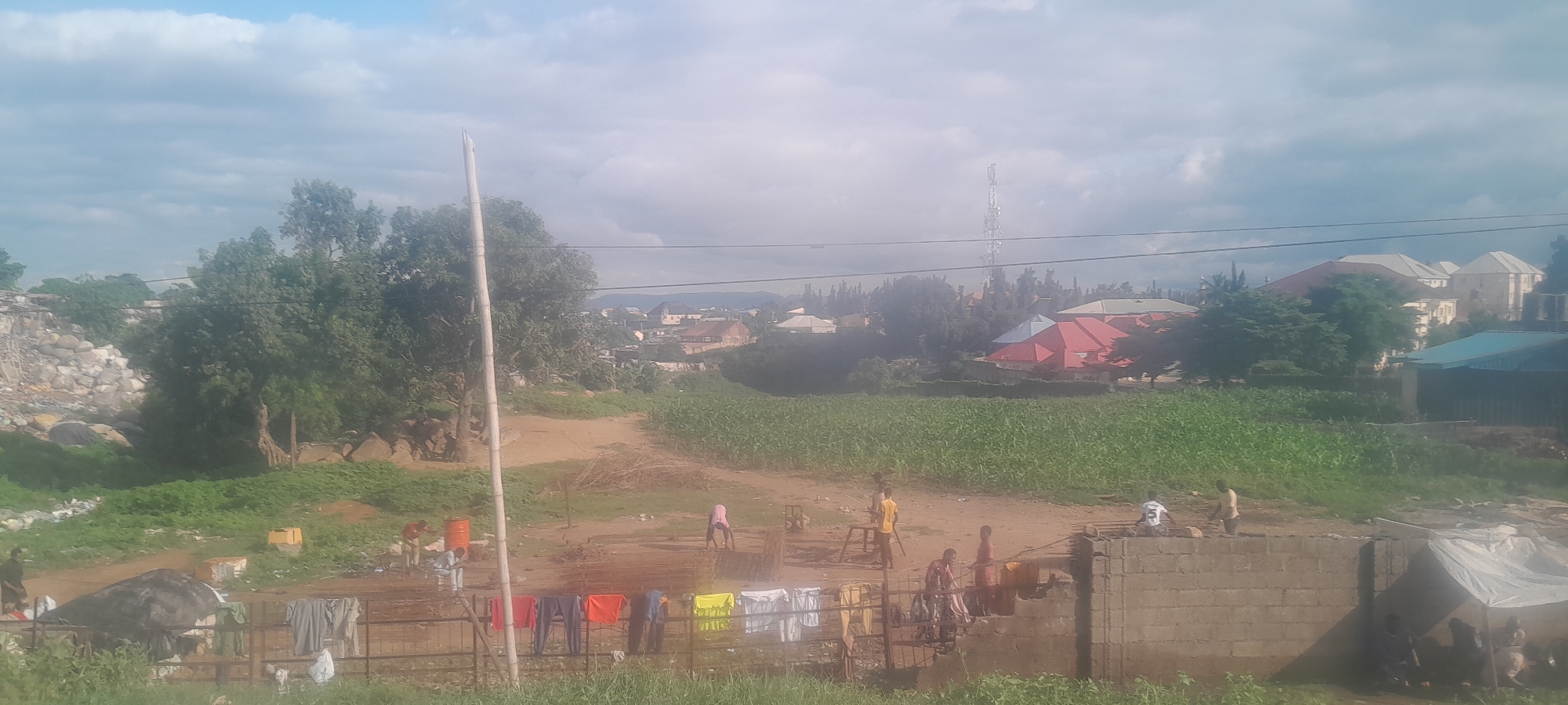 The image depicts a scene in Nyanya, Nigeria, characterized by a mix of urban and rural elements. In the foreground, a clothesline with brightly colored laundry hangs over a fence. A partially constructed brick wall runs across the mid-ground, with several individuals visible near it. To the left, a pile of debris and rocks is present, suggesting construction or demolition activity. A large field of green crops, likely corn, occupies the central portion of the image, extending towards the background. Various residential buildings, some with red roofs, and a prominent telecommunications tower are visible in the distance, indicating a settled urban area beyond the agricultural land. The sky is partly cloudy. The overall impression is one of an active, developing area where everyday life, including domestic chores like drying laundry, coexists with ongoing construction and agriculture.