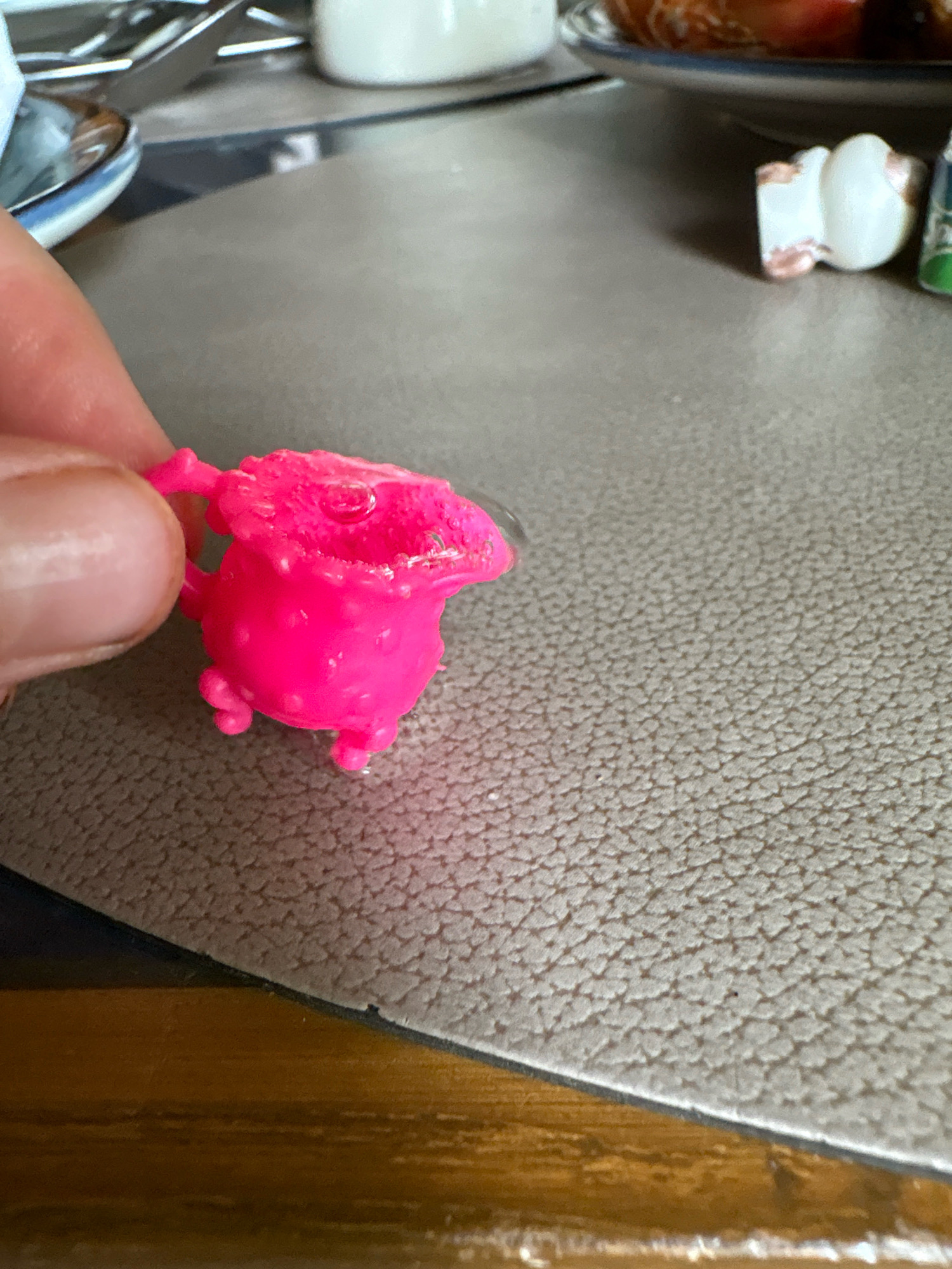 A close-up, overhead shot shows a person's thumb and index finger holding a small, bright pink, plastic toy cauldron. The cauldron has a detailed, bubbled texture and a slightly melted or distorted opening. It rests on a textured, gray surface that appears to be a placemat or table runner. In the background, out of focus, a blurred dish with some food is visible, as well as a white object and a sliver of what looks like a small figurine. The scene is illuminated by artificial light, casting subtle shadows. The location is not discernable from the provided image.