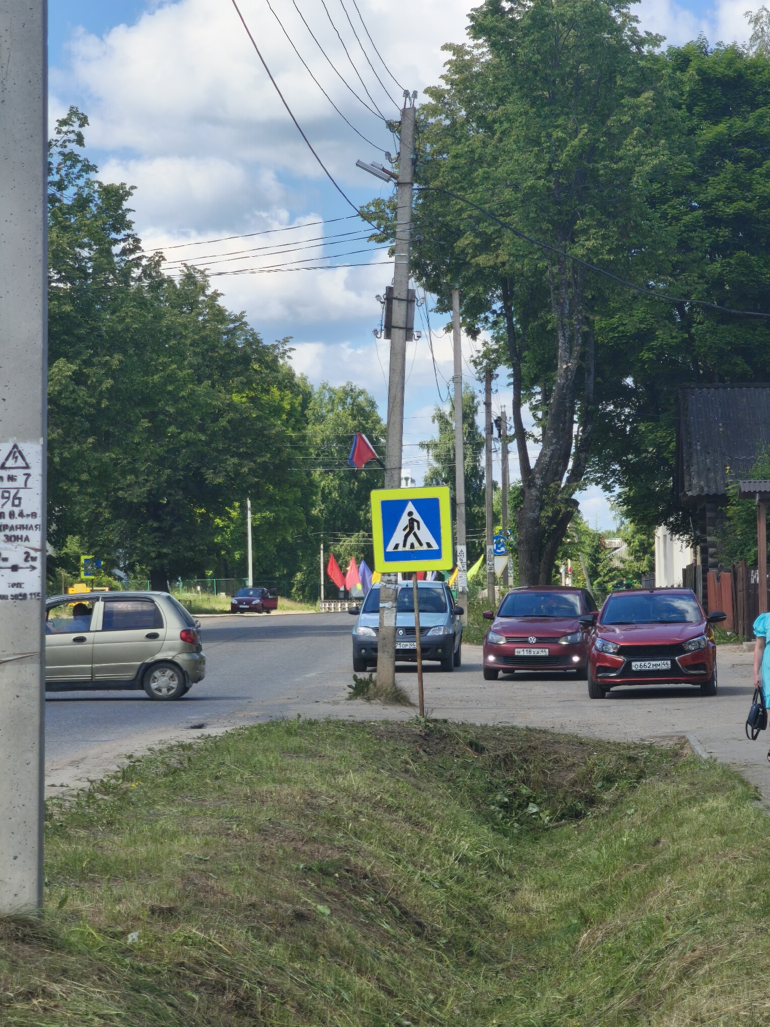 The image depicts a street scene in Kady, Russia. A concrete utility pole stands on the left foreground, with a sign attached showing a lightning bolt symbol and Cyrillic text. To the right of the pole, a light-colored sedan is parked on the side of the road. Beyond the sedan, a two-lane asphalt road stretches into the distance, with several cars driving on it. One of these cars is a red sedan with license plate "0662MM 44". In the background, tall green trees line the sides of the road, and a few red flags are visible. A pedestrian crossing sign is prominently displayed on the right side of the road, with a blue square background and a white pedestrian silhouette. The sky is partly cloudy with white clouds against a blue background. The overall environment appears to be a semi-rural or suburban area.