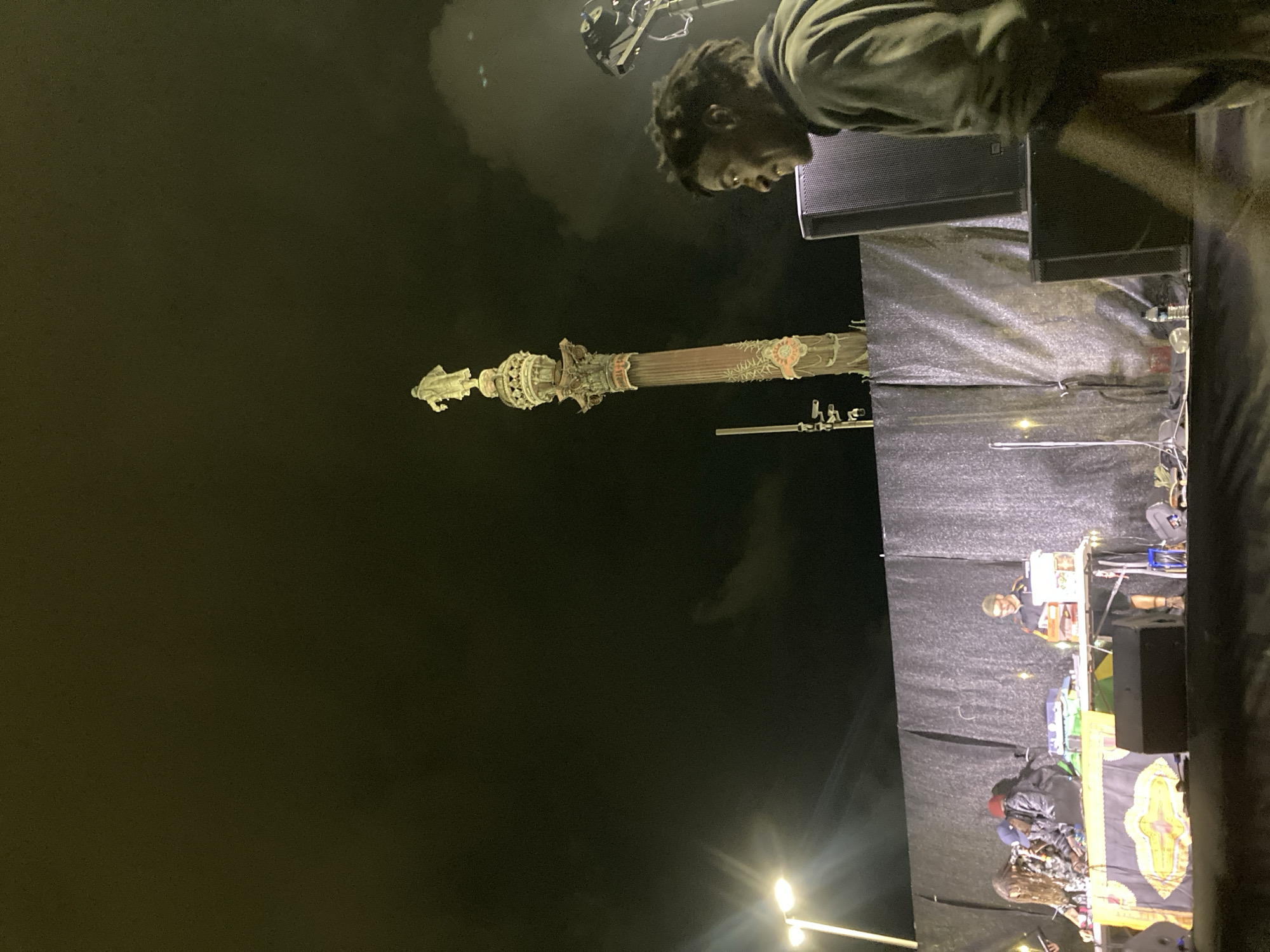 At night, in what appears to be the city of Unknown, Unknown, the Christopher Columbus monument stands in the distance against a dark sky. In the foreground, an event is underway on an elevated stage. Multiple people are positioned on the stage; one is observed standing near a speaker, looking downwards. Audio equipment and other gear are apparent on the platform. Lights are visible in the lower portion of the frame.
