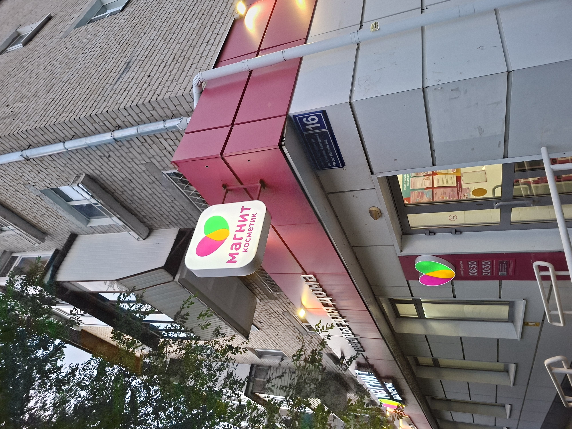 The image shows the exterior of a "Magnit Kosmetik" store, a Russian cosmetics and household goods retailer, located at Stepan Khalturin Street, building 16, in Kazan, Russia. The store's facade features a prominent red and white color scheme with illuminated signage. A large white sign with the "Magnit Kosmetik" logo, featuring a heart with colorful dots, is mounted above the entrance. Below this, another sign displays "Косметика для дома" (Cosmetics for the home). A street sign on the building indicates "ул. Степана Халтурина" (Stepan Khalturin Street) and the number "16". The store has large glass doors and windows, and information posters are visible inside. A drainage pipe runs down the side of the brick building. Trees with green foliage partially obscure the upper portion of the building. The lighting suggests it may be late afternoon or early evening.