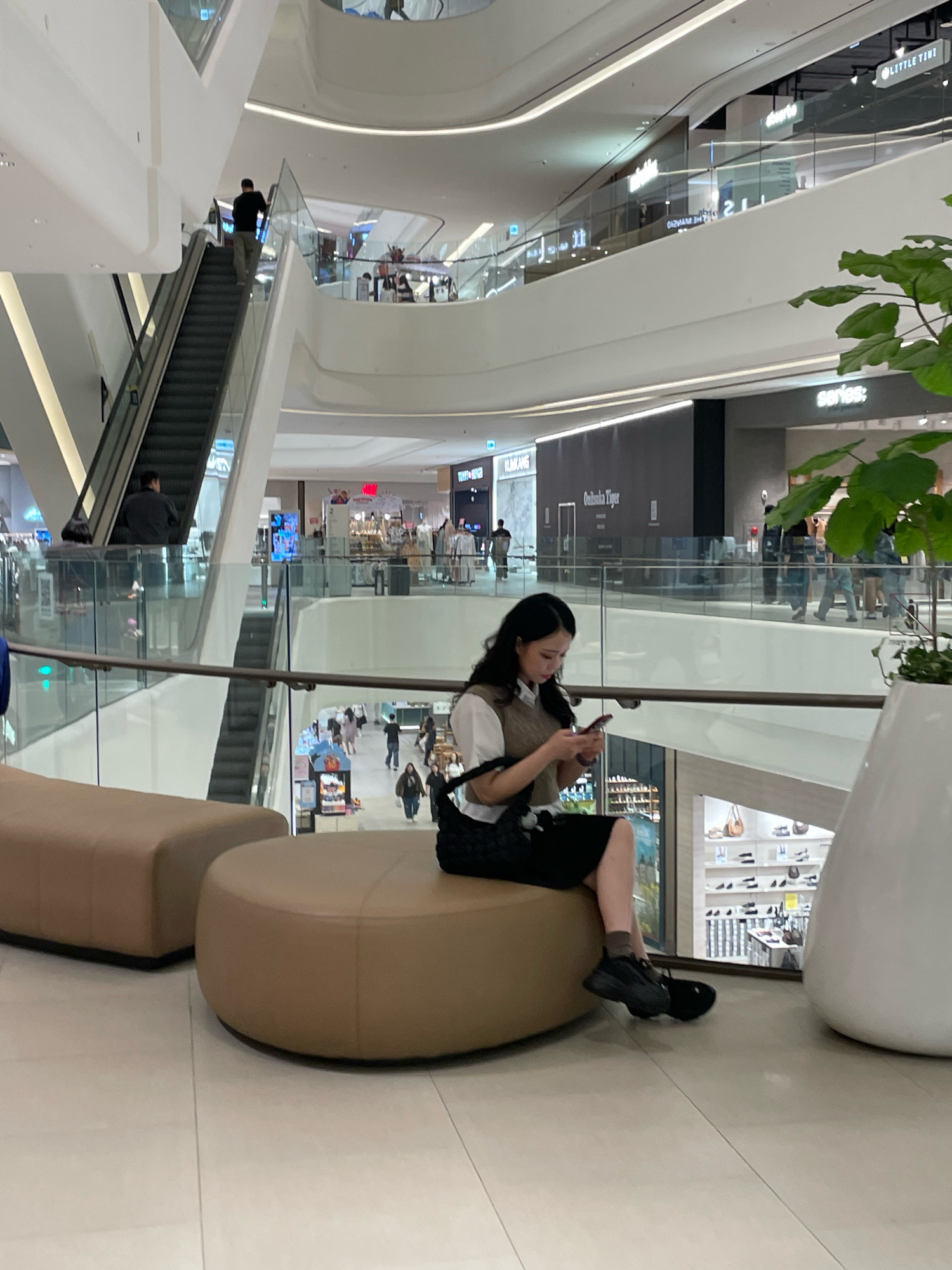 The image shows a multi-level shopping mall interior in Suwon-si, South Korea. In the foreground, a young woman with dark hair is seated on a rounded, tan ottoman, looking at her mobile phone. She is wearing a white shirt, a brown vest, a dark skirt, and black sneakers. To her left is a rectangular, tan ottoman. A plant in a large white planter is visible to the right.
In the background, a modern architectural design features curved white walls and glass railings. An escalator is visible on the left, with a person ascending it. Other shoppers are seen on different levels of the mall, browsing shops and moving around. Storefronts with signage, including "Onitsuka Tiger," are visible. The overall lighting suggests an indoor environment with artificial illumination.
