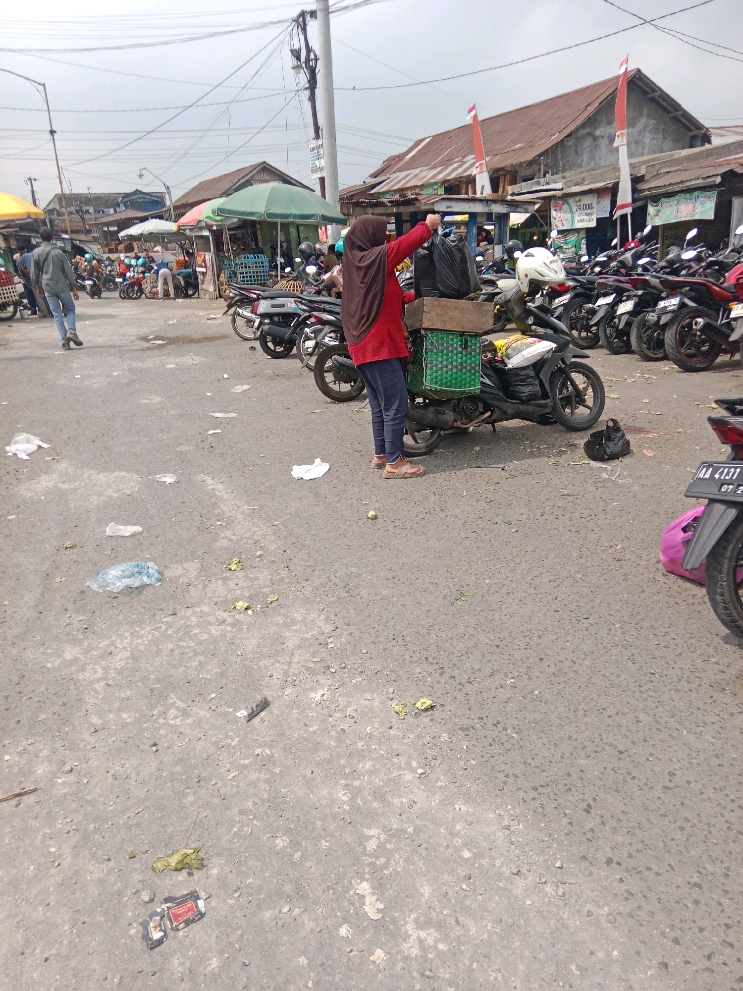 In the city of Kertek, Indonesia, a street market scene unfolds under a cloudy sky. A woman wearing a brown hijab and a red shirt is standing next to a motorcycle, holding two black plastic bags. The motorcycle is equipped with a green basket attached to the back, containing produce. Several other motorcycles are parked nearby, some with helmets on them. Various market stalls with green and white umbrellas are visible in the background, as well as buildings with rusted roofs. The ground is paved with asphalt and littered with debris, including plastic bags and food scraps. A person in a gray shirt and blue jeans is walking away from the camera on the left side of the image. Another person is partially visible in the background, near the market stalls. The overall impression is of a busy, outdoor market environment.