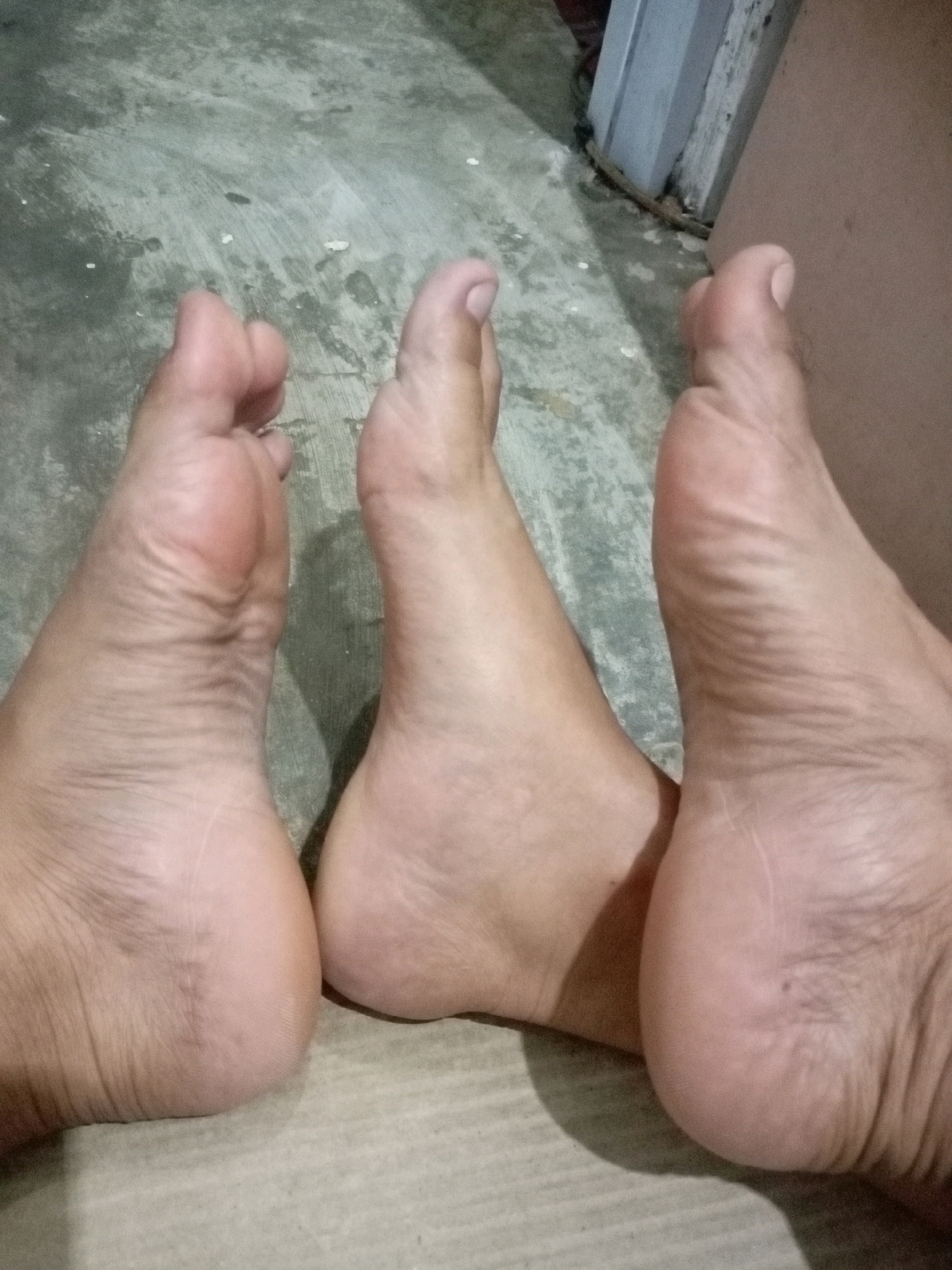 Three human feet are visible, positioned side-by-side on a concrete floor. The feet are bare and appear to be from at least two different individuals, judging by the variations in skin tone and texture. The floor is a neutral gray with some lighter and darker patches, suggesting it is an interior surface. To the right of the feet, a portion of a doorway and a wall can be observed. The lighting appears to be artificial, casting subtle shadows. The image is a close-up shot, focusing solely on the feet and immediate surroundings. The specific location of Batam, Indonesia, is not visually discernible from the image itself; this information is external to the visual content.