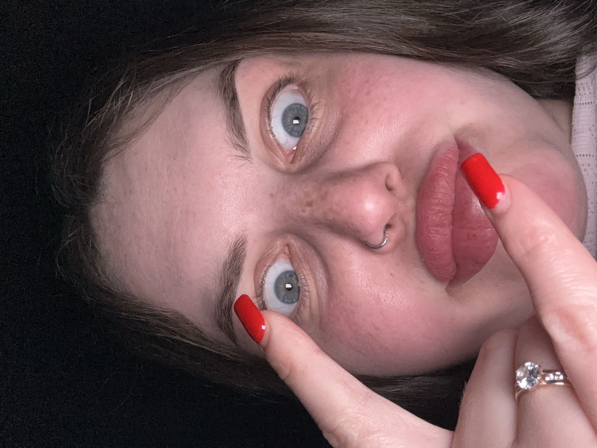 A close-up, angled portrait of a fair-skinned person with light blue eyes and brown hair is presented against a dark, indistinct background. The person's face features freckles and a nose ring. Their lips are full and colored with a muted rose shade. The subject's right index finger, adorned with red nail polish, is positioned near their eye. A gold ring with a large, clear stone is visible on the same hand. The subject's expression is one of slight surprise or alertness. The image does not suggest or contain any information pertaining to the location of Chornomorskoe, Ukraine.