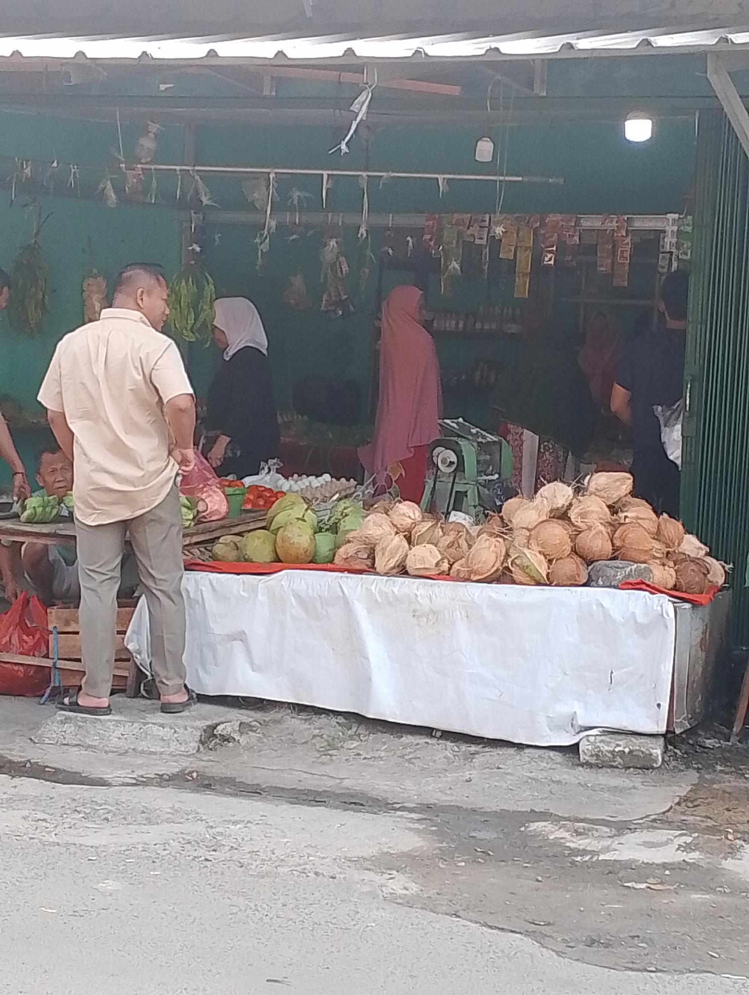 This is a medium shot, eye-level, outdoor photograph taken in a street market in Bogor, Indonesia. The foreground shows a paved area with some puddles of water. In the middle ground, a man wearing a light brown short-sleeved collared shirt and khaki pants stands facing away from the camera, looking at a market stall. A woman in a black hijab and dark clothing stands behind the stall, and another person wearing a pink hijab is visible in the background. The market stall features a table covered with a white cloth, displaying a large quantity of coconuts and some other fruits and vegetables. The background shows the interior of the market stall with shelves stocked with goods. A green metal machine is also visible in the stall. The overall scene depicts a typical market environment in Bogor.