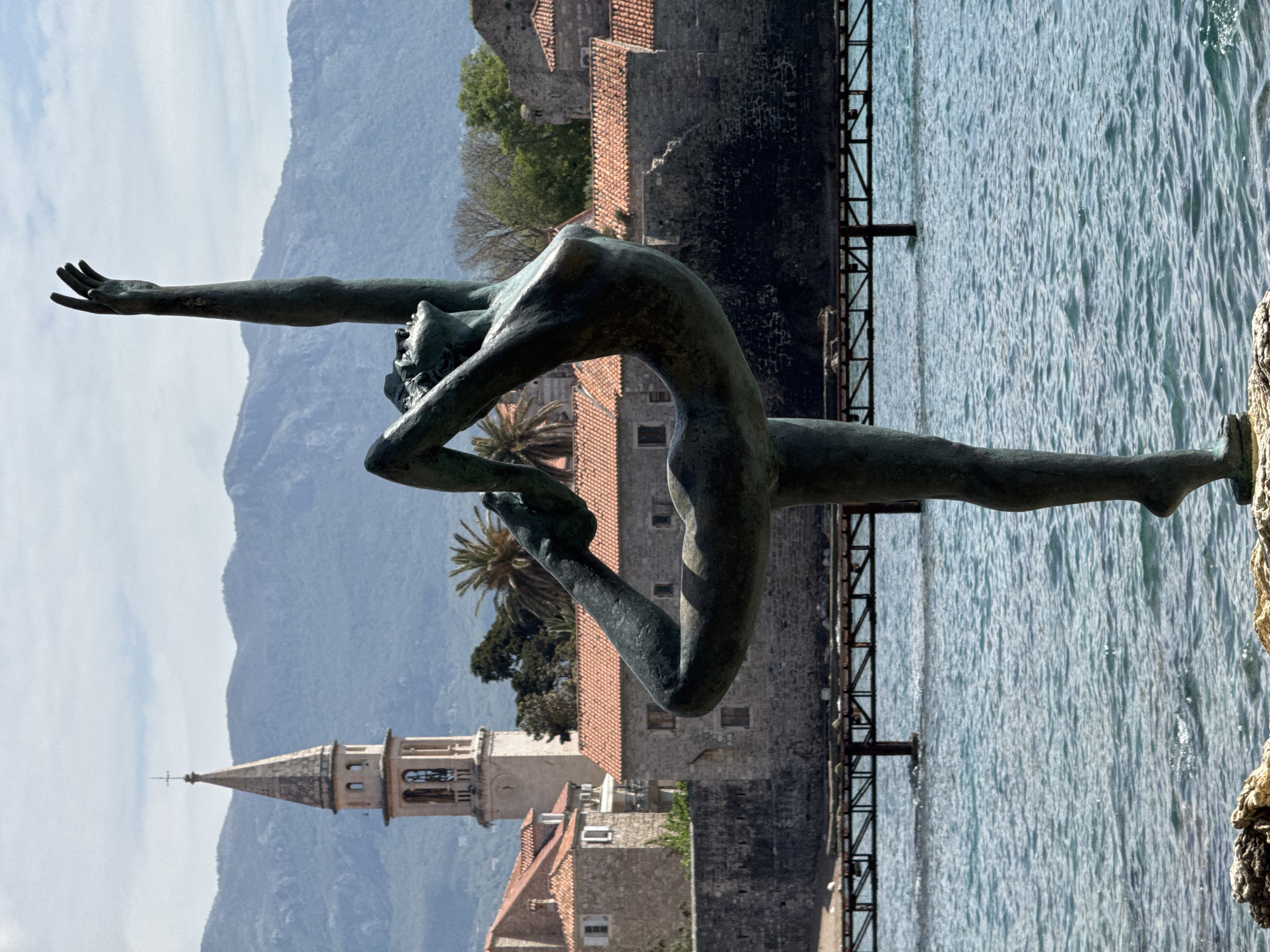 The image features the "Ballerina of Budva" statue, a bronze sculpture of a female figure in a contorted yoga pose, located in Budva, Montenegro. The statue is positioned on a rock overlooking the Adriatic Sea. Behind the statue are the old town walls and buildings of Budva, including a prominent church tower. In the background, mountains rise under a partly cloudy sky. A metal railing is visible along the water's edge. The water is turquoise in color and has small ripples.