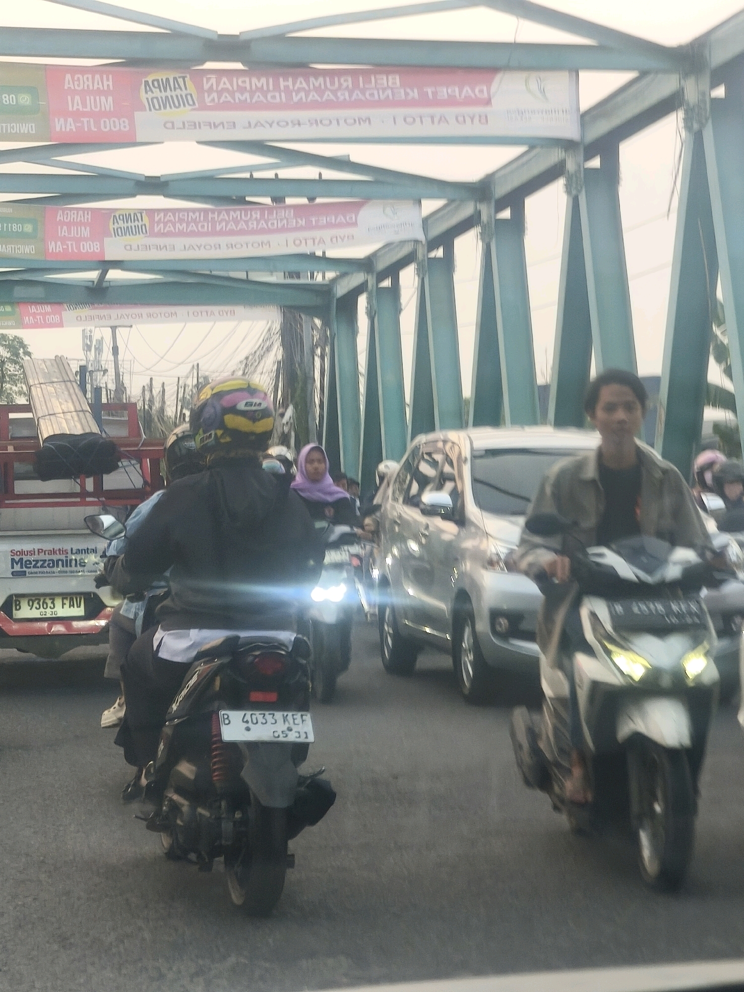 The image depicts a traffic scene on a bridge in Karangsatria, Indonesia. The bridge is supported by green metal girders. Several motorcycles and a silver SUV are present. In the foreground, a person wearing a helmet rides a black motorcycle with a license plate that reads "B 4033 KEF 05 21". To the right, another person on a white and black motorcycle is visible. Behind the SUV, a person in a purple headscarf is on a motorcycle. A white truck with the text "Solusi Praktis Lantai Mezzanine" is also in the scene. Banners with Indonesian text are hanging from the bridge structure. The overall lighting suggests it is daytime.
