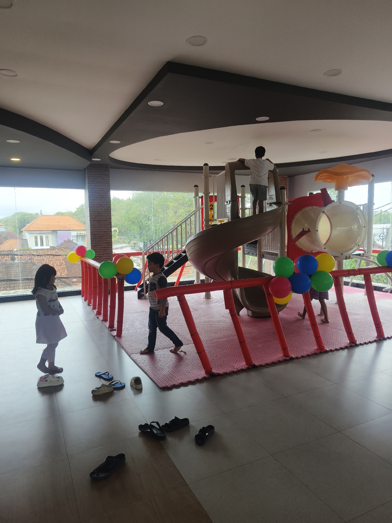 In Denpasar, Indonesia, an indoor children's playground is depicted. The scene features a large, multi-level play structure with a spiral slide and tube slides, made primarily of plastic and metal in white, brown, and red. Several children are present: a young girl in a white dress stands near a railing decorated with colorful balloons (yellow, blue, green, and red), while a boy in a striped shirt and dark pants walks across a red mat. Another individual, possibly an older child or adult, stands at the top of the play structure, facing away from the camera. The playground is situated within a room with a high, curved ceiling and large windows that offer a view of a residential area with tiled roofs and lush greenery. Scattered on the tiled floor are various pairs of children's footwear, including sandals and flip-flops. The lighting appears to be a mix of natural light from the windows and overhead artificial lighting.