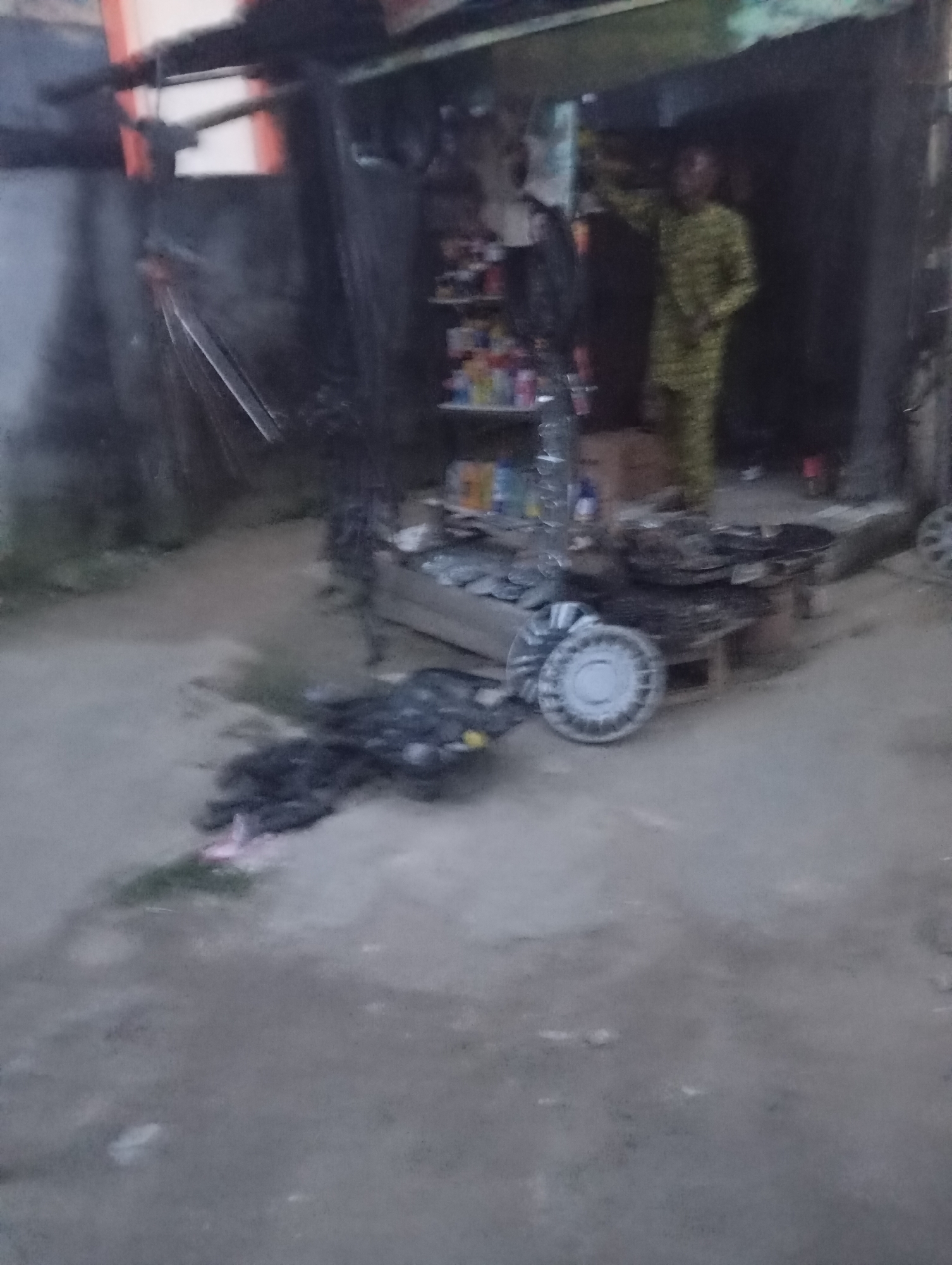 This is a medium shot, low-angle, blurry photograph taken at dusk or dawn, depicting an outdoor scene in Port Harcourt, Nigeria. The foreground shows a dusty, uneven ground with a pile of black garbage bags and some discarded items. In the middle ground, there is a wooden stall with shelves displaying various items including bottles, cans, and boxes. A person, possibly a man, dressed in a yellow patterned garment is standing in the stall, with their right arm raised. To the left of the stall, there are several metal objects, including what appears to be a car hubcap. In the background, there is a partially visible building structure. The overall impression is of a roadside stall or informal market.