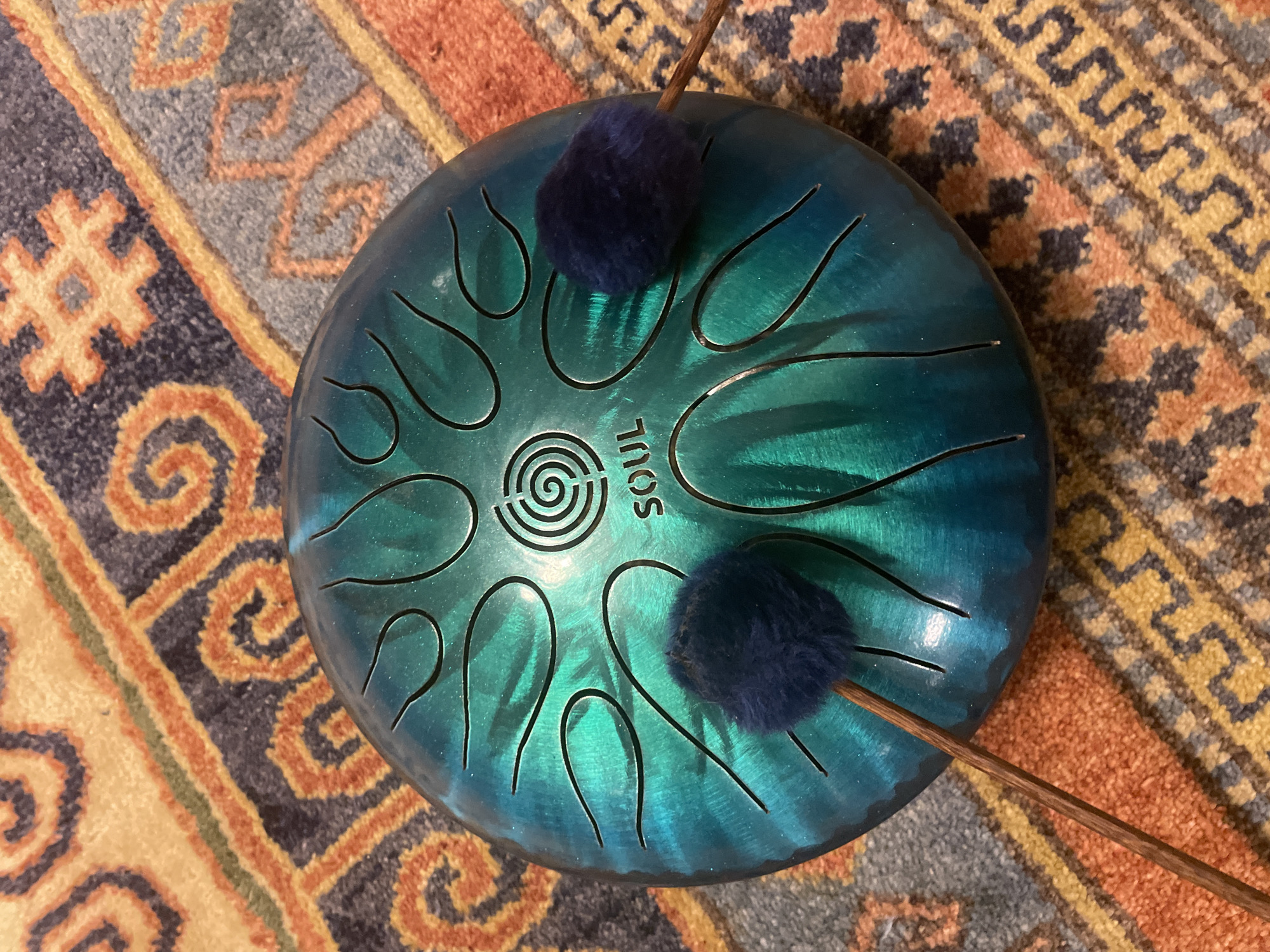 The image shows a teal-colored steel tongue drum placed on a rug with a geometric pattern. Two mallets with fuzzy blue heads rest on the drum. The drum has an engraved spiral symbol in the center with the word "SOUL" below it. The rug features a repeating pattern of orange, blue, and yellow motifs. The scene is indoors, likely in a home environment in Neoi Epivates, Greece.