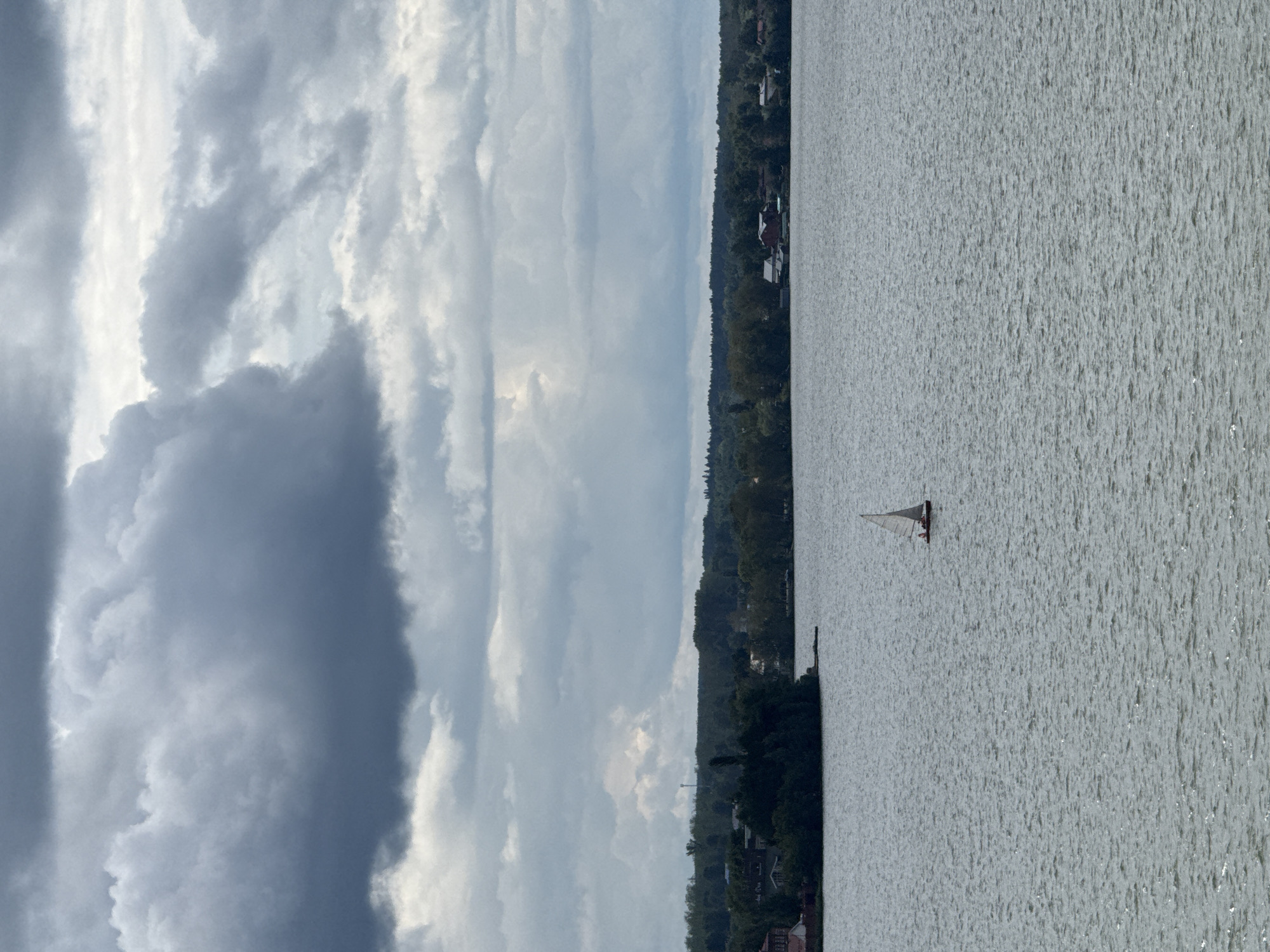 The image shows a wide body of water, likely a lake or river, under a heavily clouded sky. A small sailboat with a white sail and a red hull is visible in the middle of the water. In the background, a treeline of dense green forest stretches across the horizon, with some scattered buildings of a village or town visible nestled amongst the trees. The sky is filled with dark, dramatic clouds, suggesting overcast or stormy weather. The water has a choppy texture, indicating wind. The scene appears to be a natural landscape with a small human presence, possibly a tranquil but moody day. It is not possible to definitively identify the location as Votkinsk, Russia, based on the provided image.