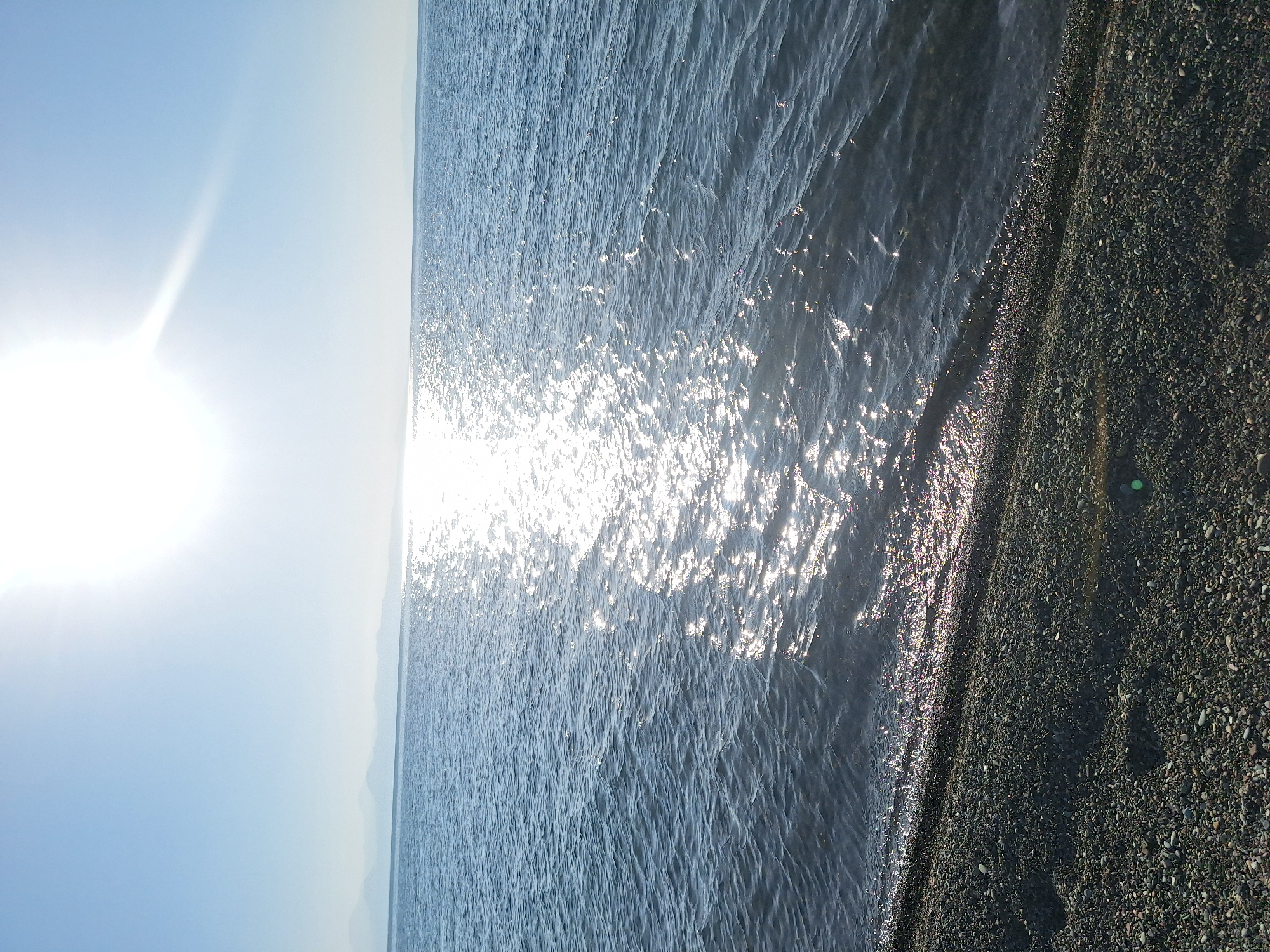 A wide shot captures a tranquil natural scene on a sunny day, likely in the vicinity of Shogakat, Armenia, a region known for its proximity to Lake Sevan. The image is dominated by the vast expanse of a body of water, presumably Lake Sevan, reflecting the bright sunlight. The water is calm, with gentle ripples disturbing the surface, and a glittering path of light stretches across it, emanating from the sun. The sun itself is prominently positioned in the upper portion of the frame, high in a clear, pale blue sky, casting a bright glare. In the distance, a hazy, mountainous horizon line is visible, suggesting a scenic landscape typical of the area. The foreground of the image is a pebble beach, composed of small, dark stones that meet the edge of the water. There are no people or man-made structures visible in the scene, emphasizing the natural and undisturbed character of the environment.