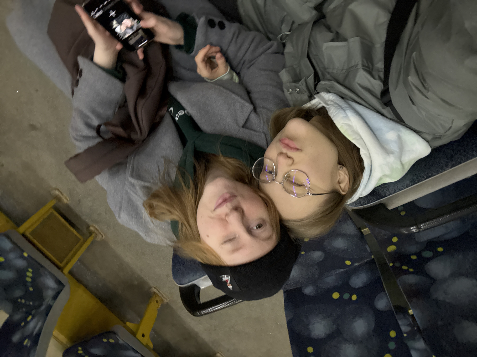 Two fair-skinned young women are lying down on a patterned blue and yellow bus seat. The woman on the left is wearing a black beanie and winking at the camera. The woman on the right is wearing round glasses. One of the women holds a cell phone. Both women wear winter coats. The interior of the bus has yellow poles and seats, indicating it is public transportation in Perm, Russia.