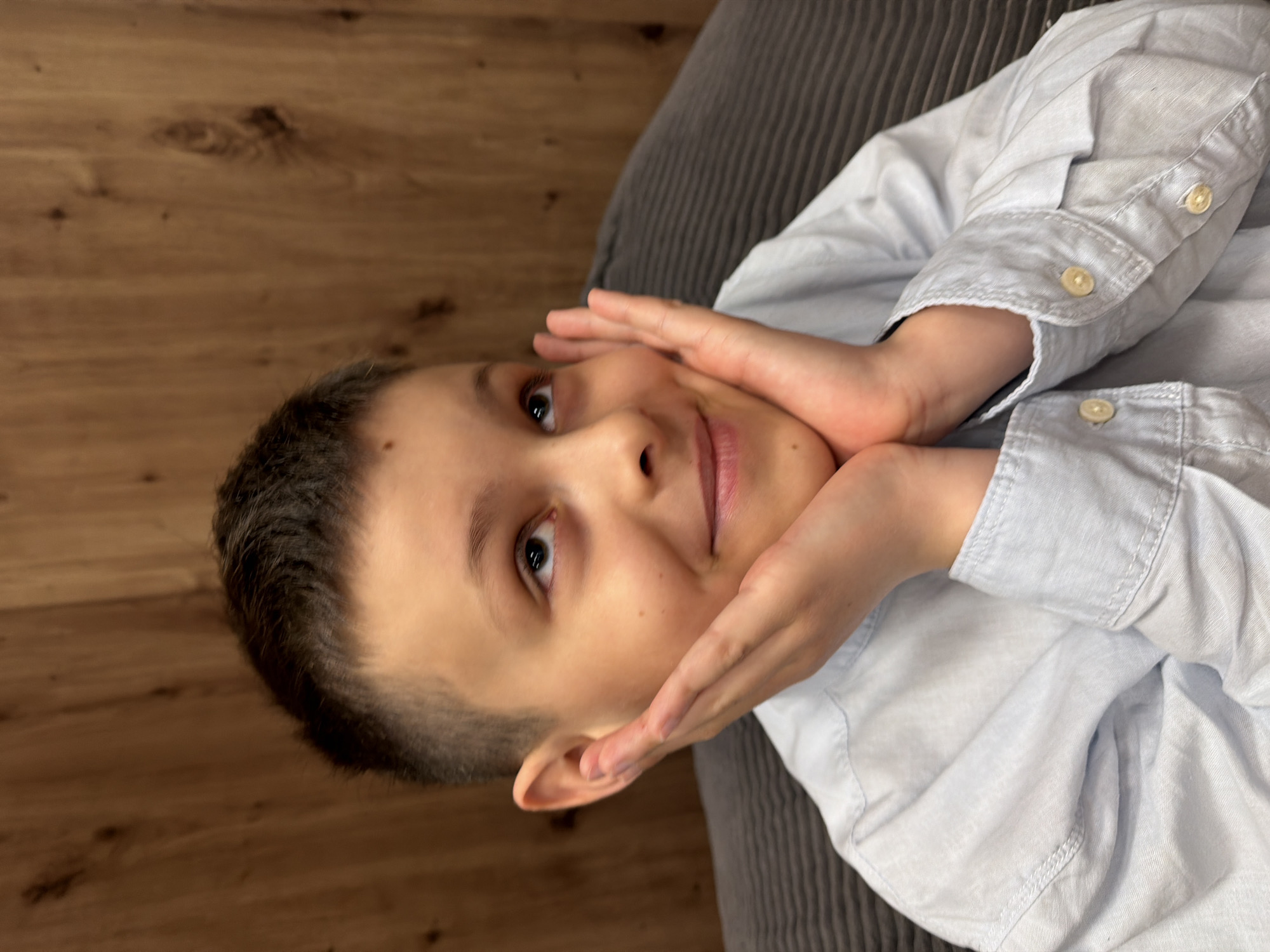 A young boy with short, dark hair and light skin is facing upward and slightly to the left. He is wearing a light blue collared shirt with light-colored buttons. His hands are positioned on either side of his face, touching his cheeks. He has a mole on his forehead and on his left cheek. The background consists of a wooden floor on the left and a gray, striped fabric surface on the right. There is no indication of the image being located in Votkinsk, Russia.