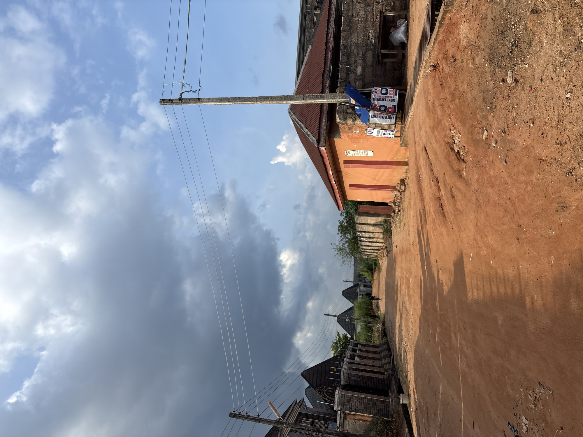 The image depicts a street scene in Obosi, Nigeria, under a sky with both bright blue patches and dark, dramatic clouds. The unpaved road, composed of reddish-orange dirt, dominates the foreground and extends into the distance. On the left side of the road, there are houses with intricate brickwork and dark, pitched roofs. Further down the street, several similar houses with pointed roofs are visible. On the right side of the road, an orange building with a corrugated metal roof stands out, and next to it is a rough stone wall. Near the orange building, several white boxes with red and blue branding, possibly advertisements, are propped up. A wooden shelf with a white sack is also present. A single person, silhouetted, walks away from the camera down the center of the road. Utility poles with multiple wires crisscross the scene, typical of the area's infrastructure. The overall impression is of a residential area in Obosi during a day with shifting weather conditions.