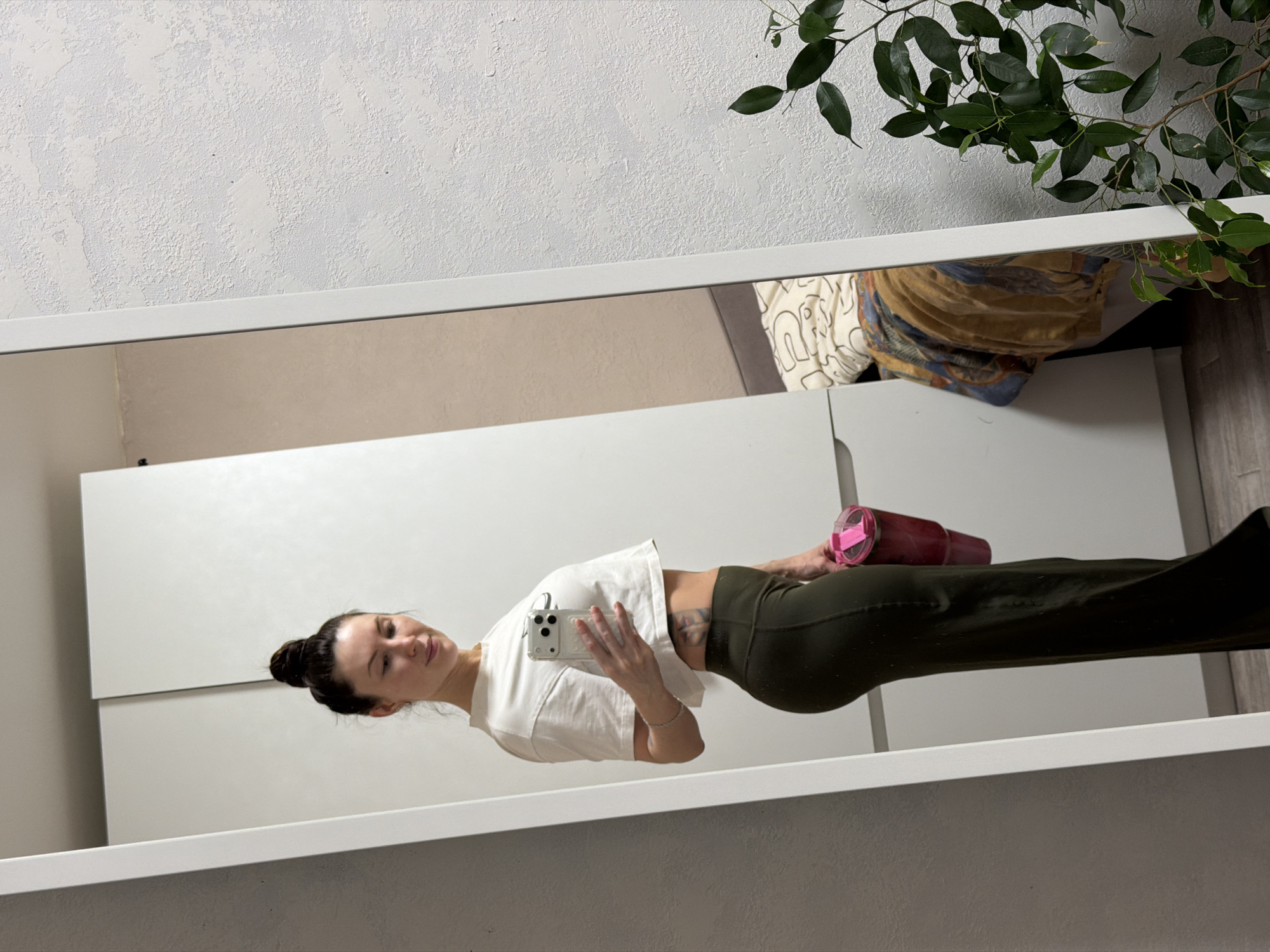 A woman is reflected in a mirror, taking a selfie with her mobile phone. She has dark hair pulled back in a bun and is wearing a white cropped t-shirt and olive-green leggings. She is holding a red and pink water bottle. The reflection also reveals part of a white cabinet or wardrobe behind her, as well as a plant with green leaves in the upper right corner of the image. The location is unclear, and there is no visual information to suggest this image was taken in Perm, Russia.