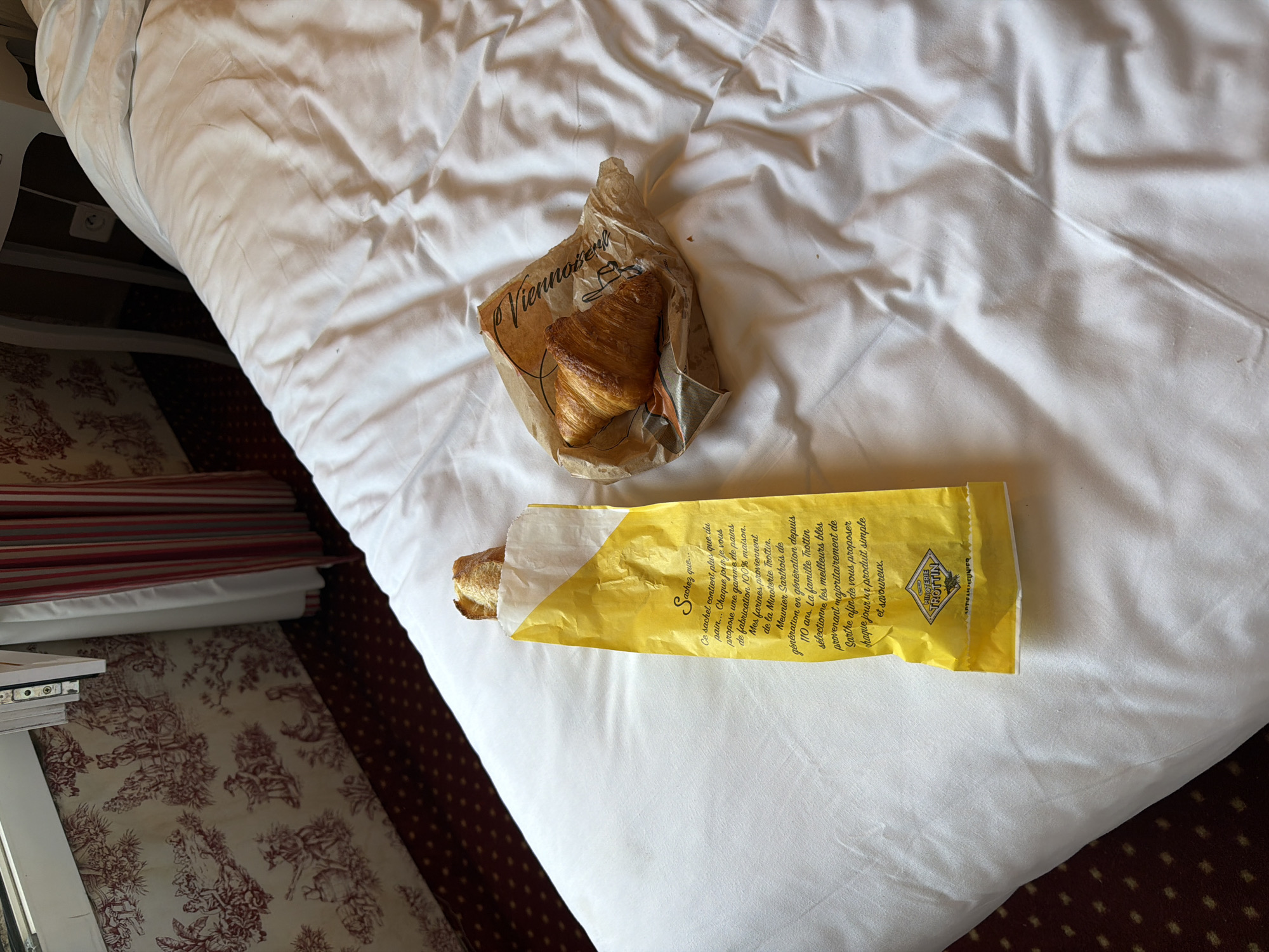 An overhead shot shows a white bed with a person's breakfast consisting of a baguette in a yellow paper bag and a croissant in a brown paper bag. The bed is covered with a white comforter that has many folds and wrinkles. The baguette bag has text written on it in French. The croissant is golden brown and flaky. The background shows a wall with red and white wallpaper and a window with red and white striped curtains. The room appears to be a hotel room, possibly in Paris, France.