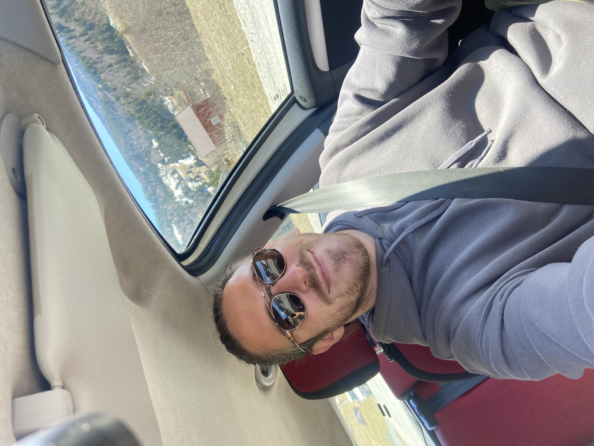 A man with a beard and sunglasses is sitting in a car, wearing a gray hoodie and a seatbelt. The car is passing through a rural landscape, possibly near Sedlačka gora, Montenegro, as seen through the window. The landscape includes bare trees and some buildings.