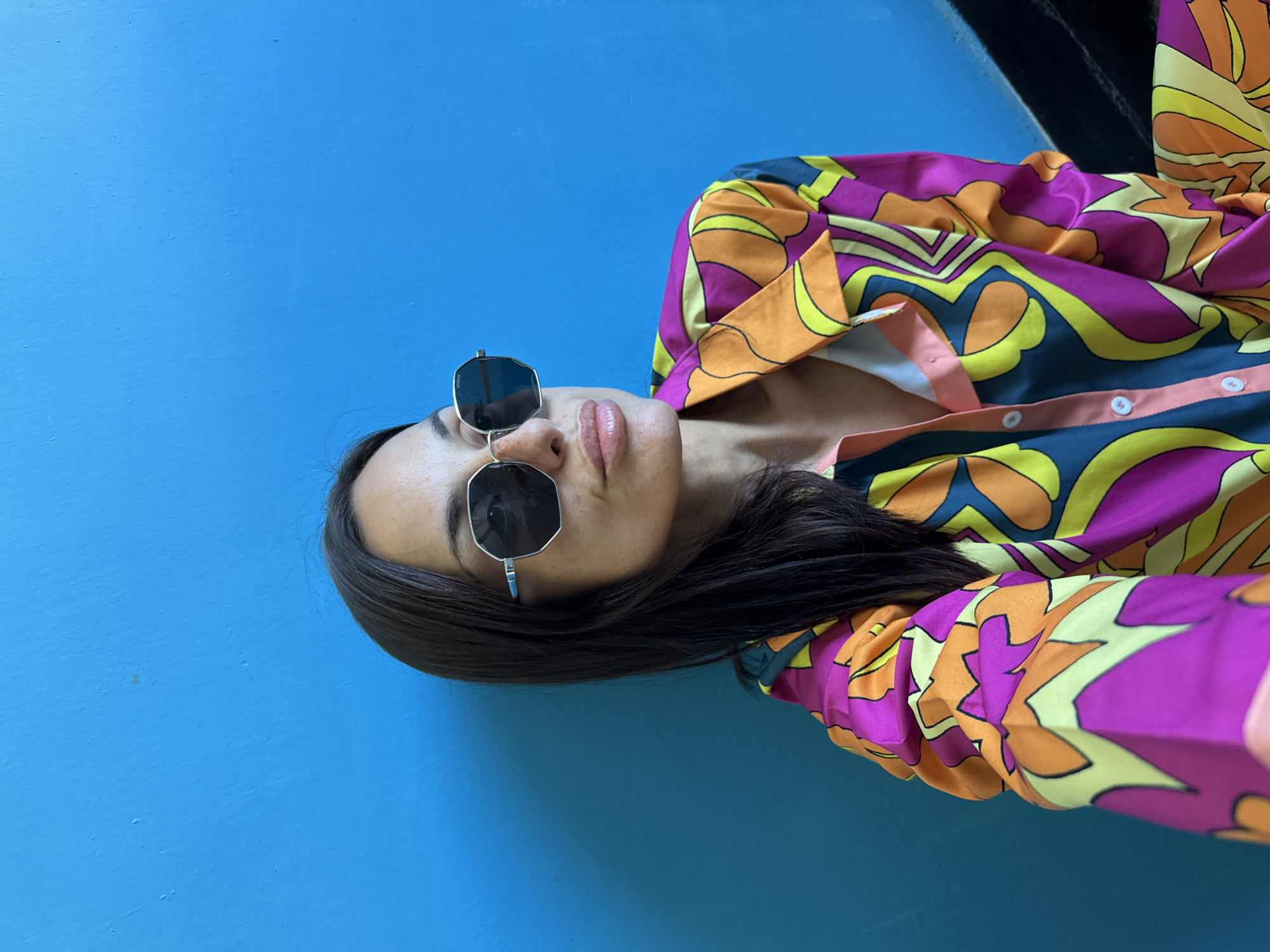 A woman with long dark hair and sunglasses is pictured against a vibrant blue wall. She is wearing a colorful, patterned shirt with shades of pink, orange, yellow, and blue. The scene does not contain any specific urban or environmental features to indicate a location within the city of Pleven, Bulgaria.