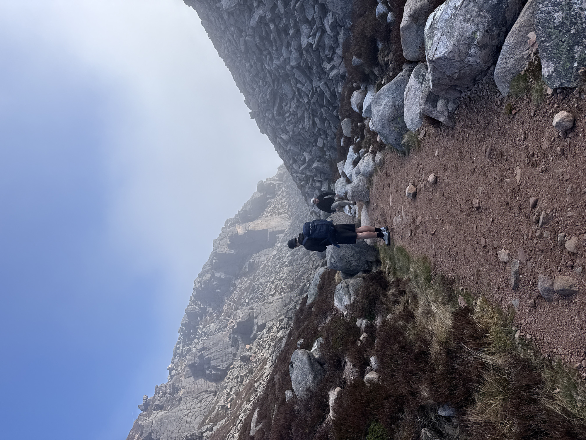 The image captures a hiking scene on a rugged mountain trail. In the foreground, a person is viewed from behind, facing away from the camera, standing on a reddish-brown dirt path. They are wearing a dark jacket, black shorts, white socks, and dark athletic shoes, and carrying a dark backpack.
The path is flanked by numerous large and small grey rocks and boulders. To the immediate right of the path, patches of dry, reddish-brown and green vegetation, possibly heather or hardy grasses, grow amidst the rocks. To the left, the terrain slopes sharply upward, revealing a massive, rocky mountain face composed of countless grey stones and crags.
Further along the path, in the mid-ground, a second person is partially visible, appearing to be walking in the same direction, but largely obscured by mist or fog. The upper left corner of the sky is clear blue, indicating daytime. However, the rest of the mountainous landscape in the mid-ground and distance is enveloped in a thick layer of white fog or low-lying clouds, creating a hazy and atmospheric environment. The scene suggests a challenging hike in a high-altitude or rugged rocky region with intermittent clear weather. No text is visible in the image.