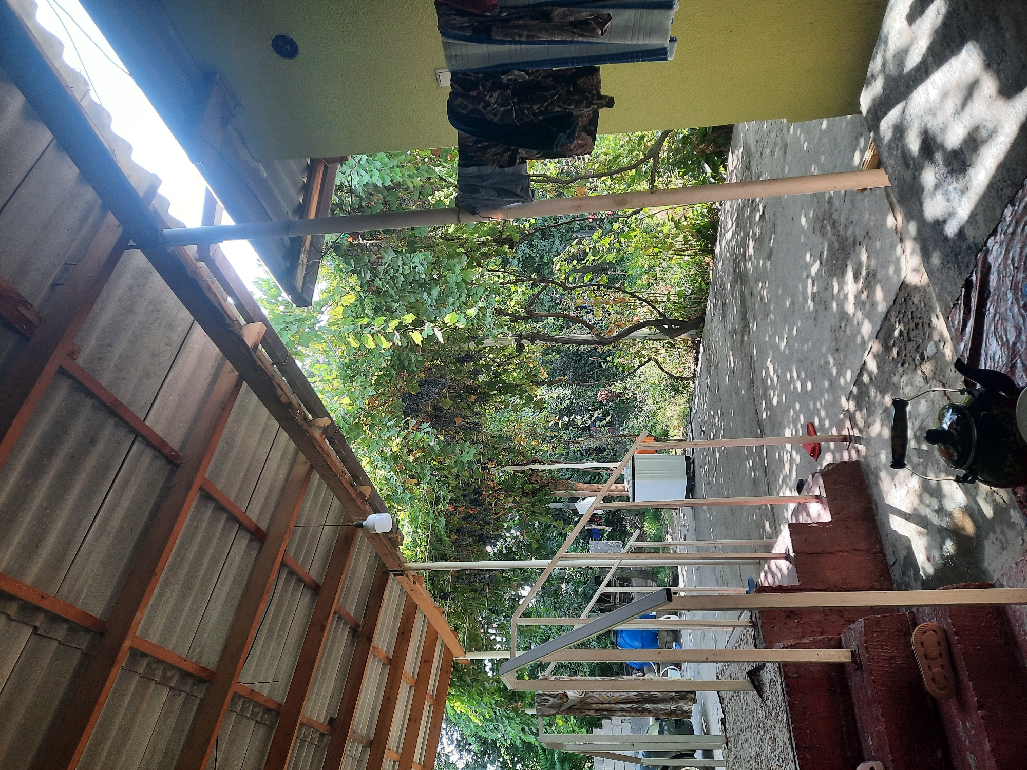 The image depicts an outdoor covered patio area in Восеабад, Tajikistan. A corrugated metal roof, supported by wooden beams, is visible overhead. A white LED light bulb hangs from a wire. To the right, a light yellow wall of a building is present, with several items of clothing hanging on a clothesline. A wooden pole supports the roof near the wall. The ground is concrete, with shadows from overhead foliage creating a dappled effect. In the foreground, a dark teapot with a patterned design sits on a surface, next to what appears to be a small cup. Red flip-flops are scattered on the ground near a set of brick steps. To the left, a metal railing leads up the steps. Behind the railing and steps, a white cylindrical object, possibly a washing machine or water tank, is situated. Beyond the patio, dense green foliage and trees are visible, with some grapevines laden with dark grapes. In the background, a blue barrel and a portion of a car are partially obscured by the vegetation and railing. The scene suggests a domestic setting in a rural or semi-rural area. There are no people visible in the image.