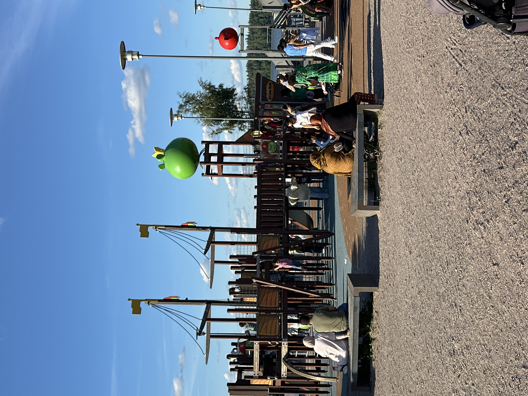 The image shows a playground on a sunny day, with people of various ages enjoying themselves. The playground features a wooden structure resembling a pirate ship, complete with masts, sails, and rigging. There are also some colorful, spherical decorations scattered around, including a large green frog-like object and a red ball. Several people are seen interacting within the playground, some sitting, some standing, and some appearing to be playing. The ground of the playground is covered in gravel, and there are benches and shaded areas for relaxation. In the background, there are trees and a body of water, suggesting the playground is situated in a scenic outdoor location. The sky is a clear blue with a few scattered clouds. The lighting indicates it is daytime, likely late afternoon given the long shadows. The overall mood seems relaxed and cheerful, with families and individuals spending leisure time outdoors.