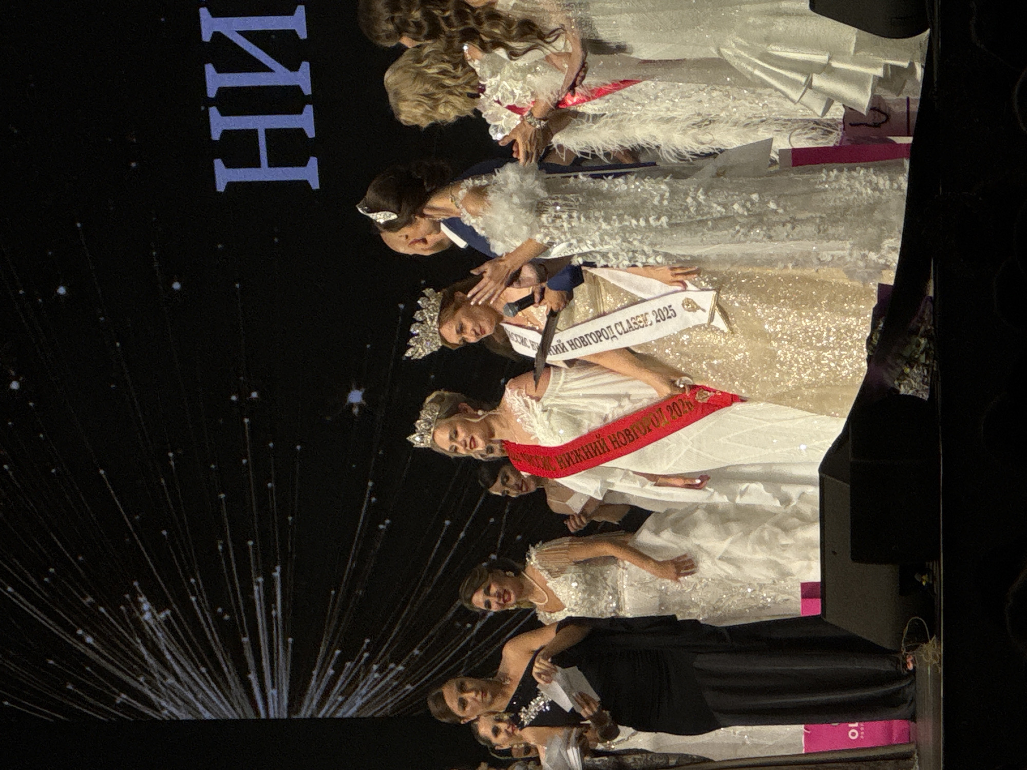 The image shows a group of women, primarily beauty pageant contestants, likely at an event in a city whose name includes "Нижний". The women are wearing gowns and sashes, with some also sporting crowns. One sash reads "НИЖНИЙ НОВГОРОД CLASSIC 2025", possibly indicating the name and year of the pageant. The image is taken from a low angle, and the background features abstract light patterns against a dark background.