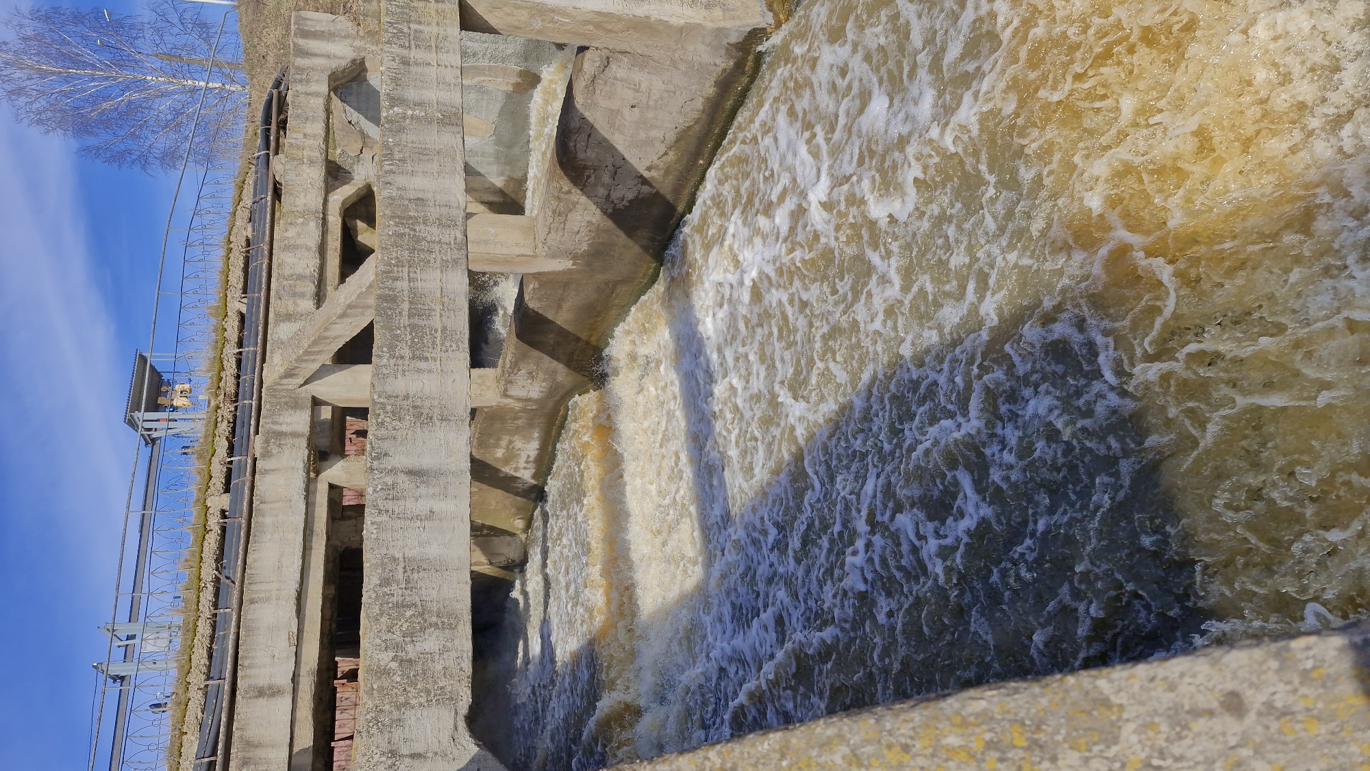 The image shows a concrete dam structure in Otchor, Russia, with water cascading over its terraced steps. The water is frothy and turbulent, its color a mix of yellow and white. To the left of the frame is the dam's vertical concrete support structure, featuring rectangular openings. A metal railing runs along the top of the structure. In the background, the blue sky and a bare tree are visible.
