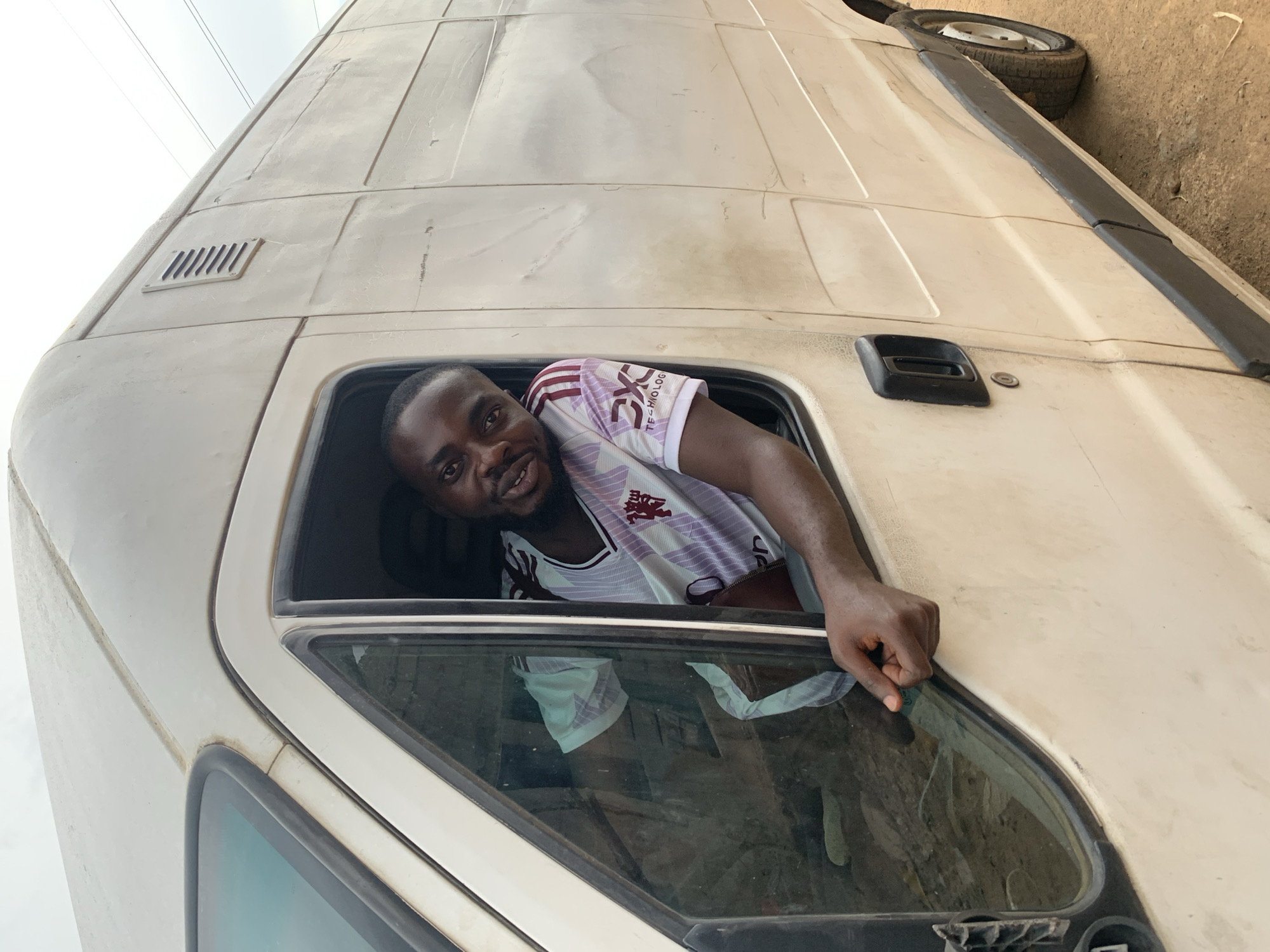 A man with a dark complexion is leaning out of the driver's side window of an old, white van. He is wearing a white and maroon jersey. The van appears to be parked on a dirt or gravel surface, with a tire visible at the top of the frame. The scene is likely in Jos, Nigeria.