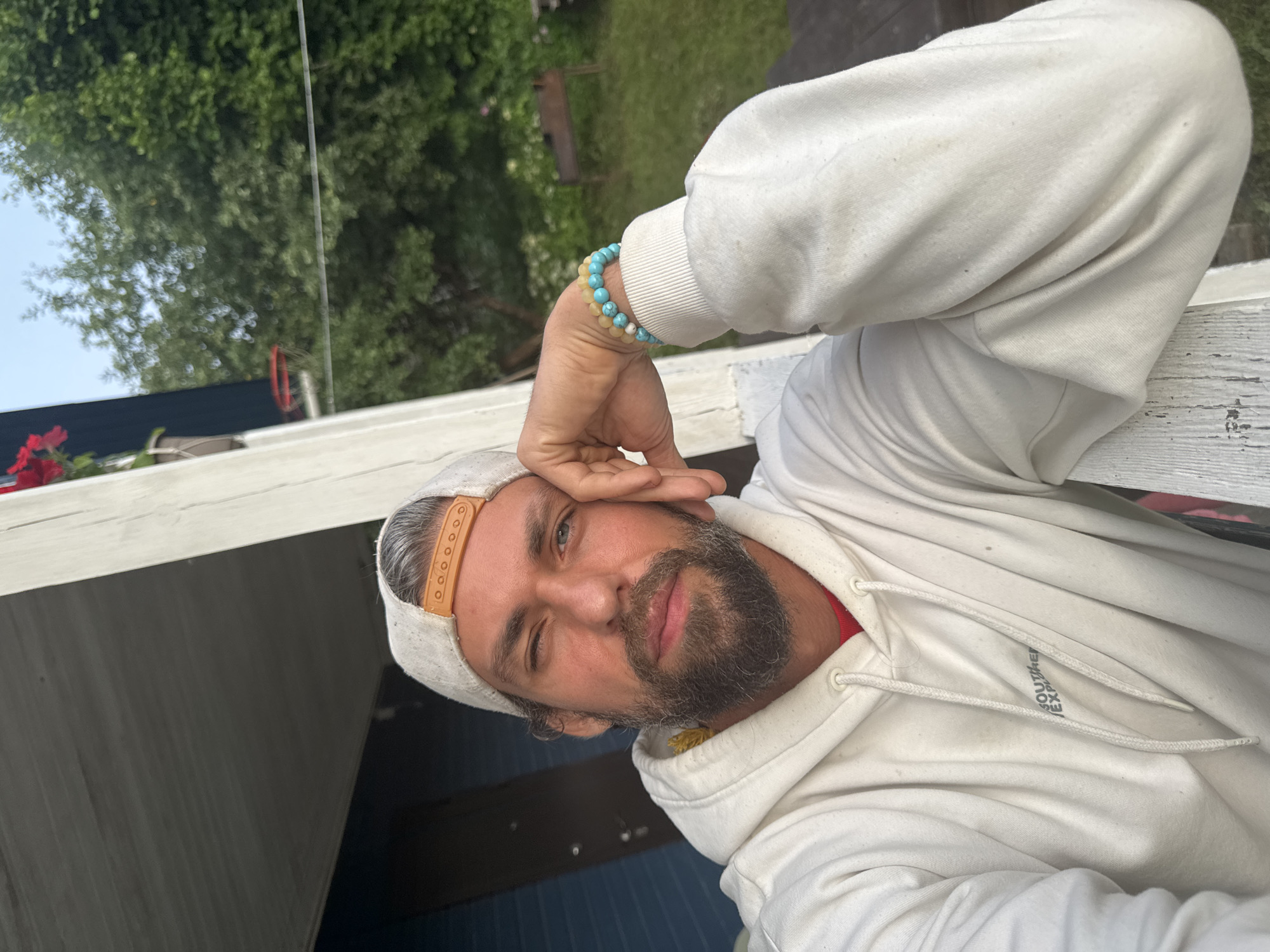 The image is a close-up, eye-level, selfie-style photograph of a man with graying hair, a beard, and blue eyes. He is wearing a light-colored baseball cap, a white hoodie over a red shirt, and a bracelet made of light blue and yellow beads on his left wrist. He is resting his head on his left hand, and his left eye is partially closed. The background appears to be an outdoor setting, possibly a yard or garden, with lush green trees and foliage. A wooden structure, possibly a porch or balcony railing, is visible in the foreground and to the right. There is no identifiable signage or landmarks to indicate the specific location.