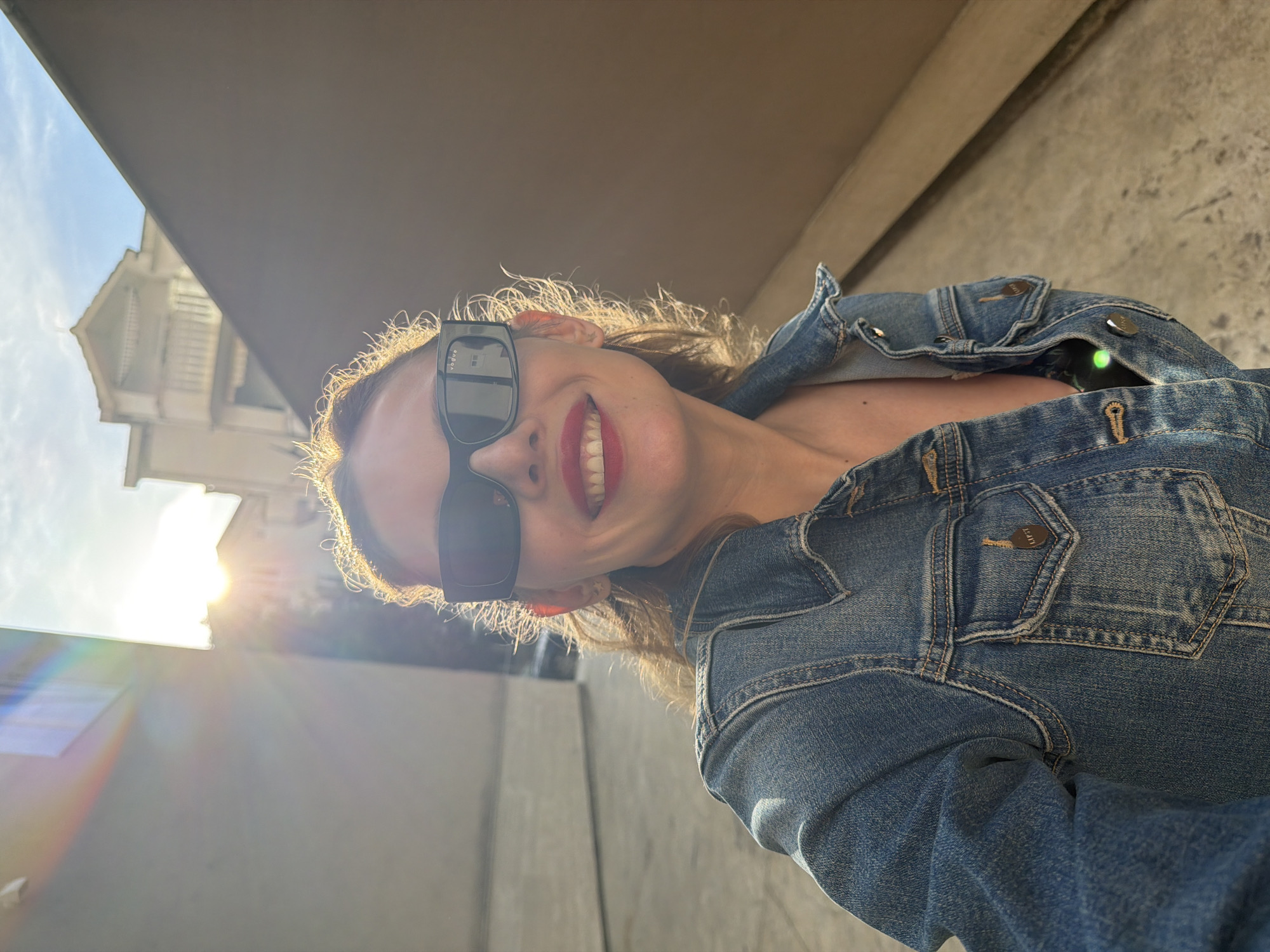 A vibrant, close-up outdoor portrait features a woman smiling broadly at the camera on a sunny day in Budva, Montenegro. She wears dark, rectangular sunglasses with "Vogue" faintly visible on the left temple arm, and bright red lipstick. Her medium-length hair is illuminated by strong backlighting, creating a radiant halo effect around her head.
She is dressed in a light blue denim jacket, with visible buttons and stitching details. The text "R.N.I." is embossed on a button near her right shoulder, and "L.P.S.Y." on a button on a back pocket. A hint of a dark garment is visible beneath the jacket. A distinct bright green light or reflection appears on her right shoulder area.
The background to the woman's left reveals part of a white building with a multi-tiered, architectural structure and railings, under a clear, bright sky. Strong sunlight, coming from the upper left, creates a prominent lens flare, indicating a bright, sunny environment, possibly late afternoon or early morning. The overall mood is joyful and confident.