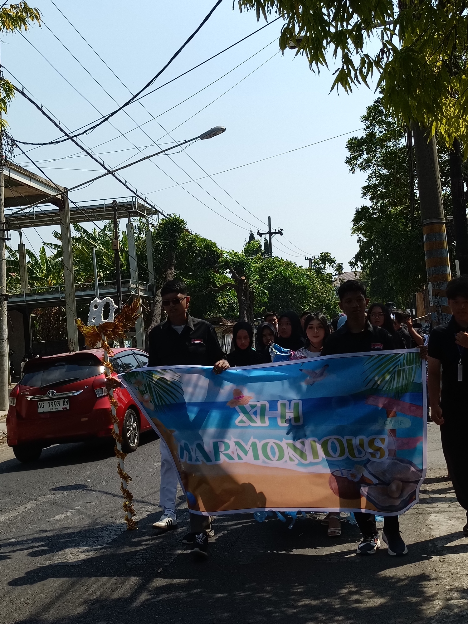 A group of young people are holding a banner that reads "XI-H HARMONIOUS" on a street in Kediri City, Indonesia. The banner features an image of a beach scene with palm trees and a large pearl. The people are dressed casually, and some are wearing black shirts. A red car is parked on the side of the road behind them. The scene appears to be a parade or a procession. The background shows trees and buildings, suggesting an urban or suburban setting. The sky is clear and blue, indicating a sunny day.