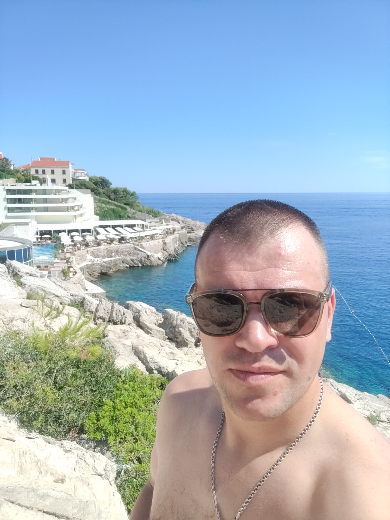 A man in sunglasses and a silver chain is taking a selfie outdoors with a resort and the ocean in the background. The man is positioned in the foreground, facing the camera. He has a shaved head and is shirtless. Behind him, a white resort building with red roofs is built into a rocky hillside overlooking a turquoise body of water. Sun loungers and umbrellas are visible on a terrace area of the resort. The sky is clear and blue.
