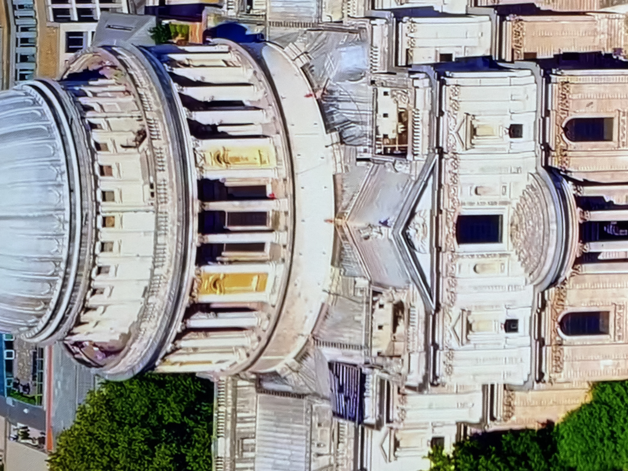 An aerial or high-angle view captures the upper sections of St Paul's Cathedral in London, United Kingdom. The prominent feature is the light grey, ribbed dome, situated atop a drum structure. This circular drum is adorned with numerous white columns and features alternating recessed windows and niches, some with yellowed interiors. A balustraded walkway runs around the base of the drum. Below the drum, the roofline of the main cathedral structure is visible, composed of light grey, possibly lead, roofing with some areas appearing textured or damaged. Further down, the intricate stone facade of the cathedral is detailed, featuring classical architectural elements such as pediments, pilasters, cornices, and arched windows. Lush green trees are visible in the lower-left and upper-left portions of the image, bordering the cathedral grounds. In the upper background, parts of other modern buildings with various rooflines and windows can be seen. No discernible people or specific actions are clearly visible.