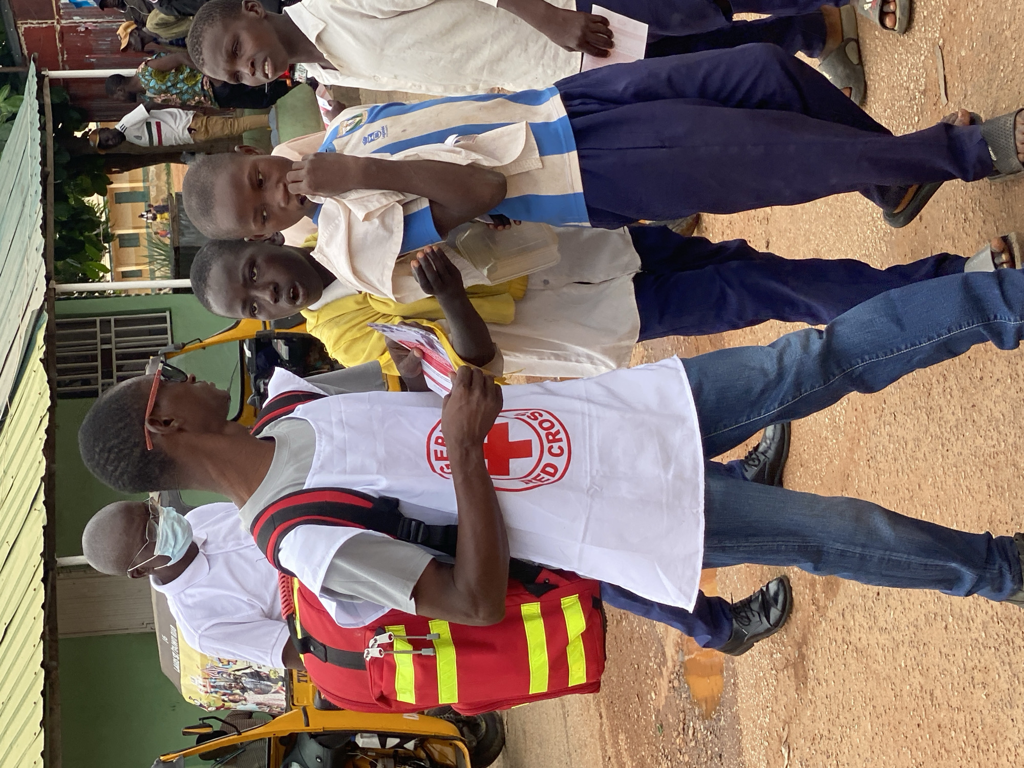 In Dadi Riba, Nigeria, on a dirt road near a building with a green corrugated metal roof, a group of people are gathered. In the foreground, a young person wearing a white vest with the Red Cross emblem walks forward, carrying a red medical bag with yellow reflective stripes on their back. They are wearing blue jeans and black shoes. To their right, two other young individuals are standing. One wears a yellow shirt and blue pants, and the other wears a blue and white striped shirt and blue pants. Further back, an adult wearing a white shirt and glasses, with a face mask, stands near a yellow tricycle. Several other individuals are visible in the background. The ground is muddy in places, suggesting recent rain.