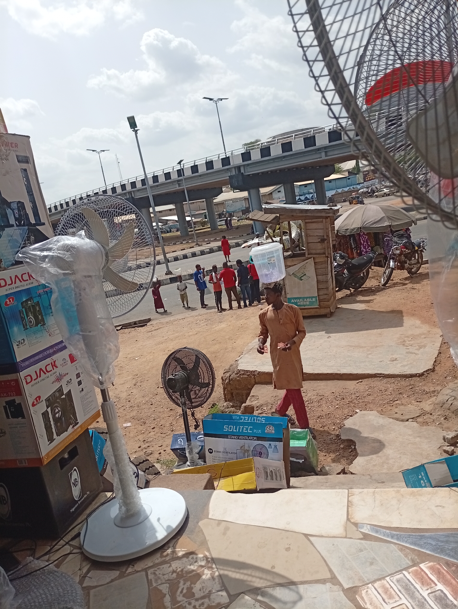 The image depicts an outdoor scene in Lokoja, Nigeria, showcasing a street-side commerce setting. In the foreground, there are several standing electric fans, some packaged and others displayed openly. Boxes of speakers from the brand "DJACK" are stacked next to the fans. A man in a brown robe and red trousers is carrying a plastic container on his head. Behind him, a small wooden kiosk-like structure is visible. In the background, there is a group of people standing near a road junction and an elevated road or bridge. Motorcycles are parked near the kiosk. The weather appears sunny with scattered clouds. The presence of "Nigerian Breweries Plc" labeled on one of the boxes suggests a possible connection to a local distributor.