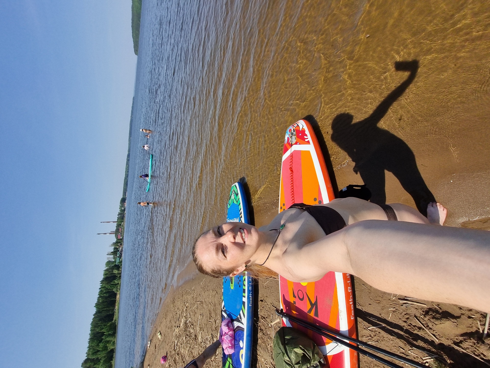 On a sunny day, a woman lies on an orange and pink paddle board on a sandy beach along a riverbank. She is fair-skinned and wearing a dark bikini. A blue paddle board is next to her. Her shadow, holding a phone, is cast on the sand and shallow water. In the background, other people are on paddle boards in the river. Across the river, there are trees and buildings, including tall industrial smokestacks which identify the location as Chaikovsky, Russia.