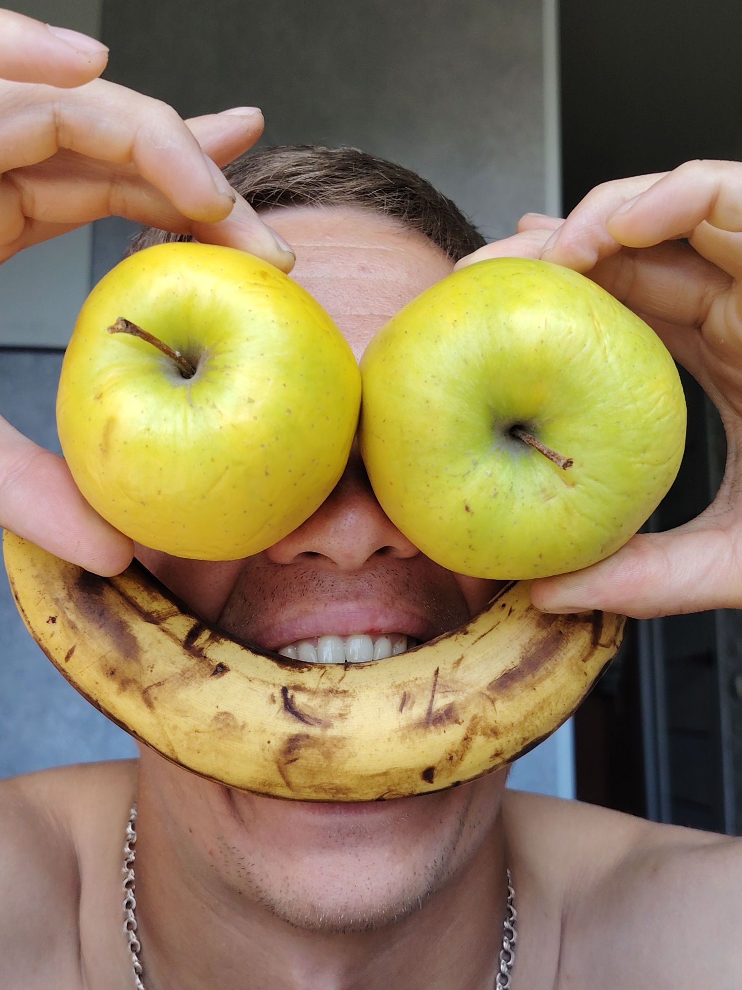 A person is holding two yellow apples in front of their eyes and a banana in front of their mouth, creating a smiley face. The person's face is partially visible, with their eyes, nose, and mouth showing. The background is blurred, but appears to be an indoor setting with a grey wall and a doorway. The image is cropped closely to the person's face and the fruit. There is no information about the location in the city of Chaikovsky, Russia.