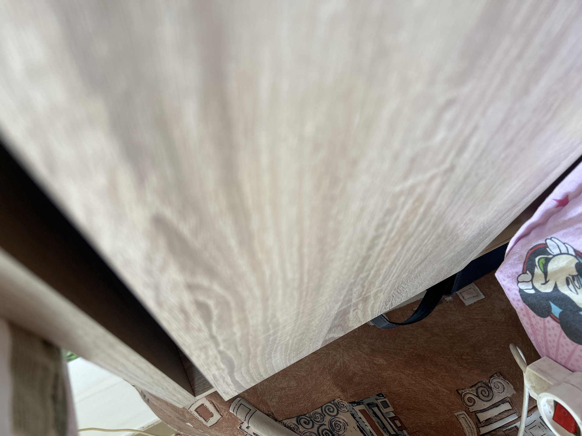 The image shows a close-up, angled view of a light-colored wooden surface, possibly a table or shelf, with a prominent wood grain pattern. The surface extends diagonally across the frame, with the upper portion out of focus and the lower portion sharper.
Below the wooden surface, a patterned brown carpet or rug is visible. On the carpet, there is a rolled-up mat with a black and white geometric design. To the right of the wooden surface, a pink fabric is draped, featuring a large image of Mickey Mouse. Part of a white electrical device with a cord is also visible on the far right. A dark strap, possibly from a bag, is visible underneath the pink fabric and on the wooden surface.
The lighting suggests an indoor setting, possibly during the day given the bright illumination on the wooden surface. The overall impression is of a casual, domestic environment. There is no visible text in the image. The scene does not depict any people or specific activity, but rather a collection of objects and surfaces.
