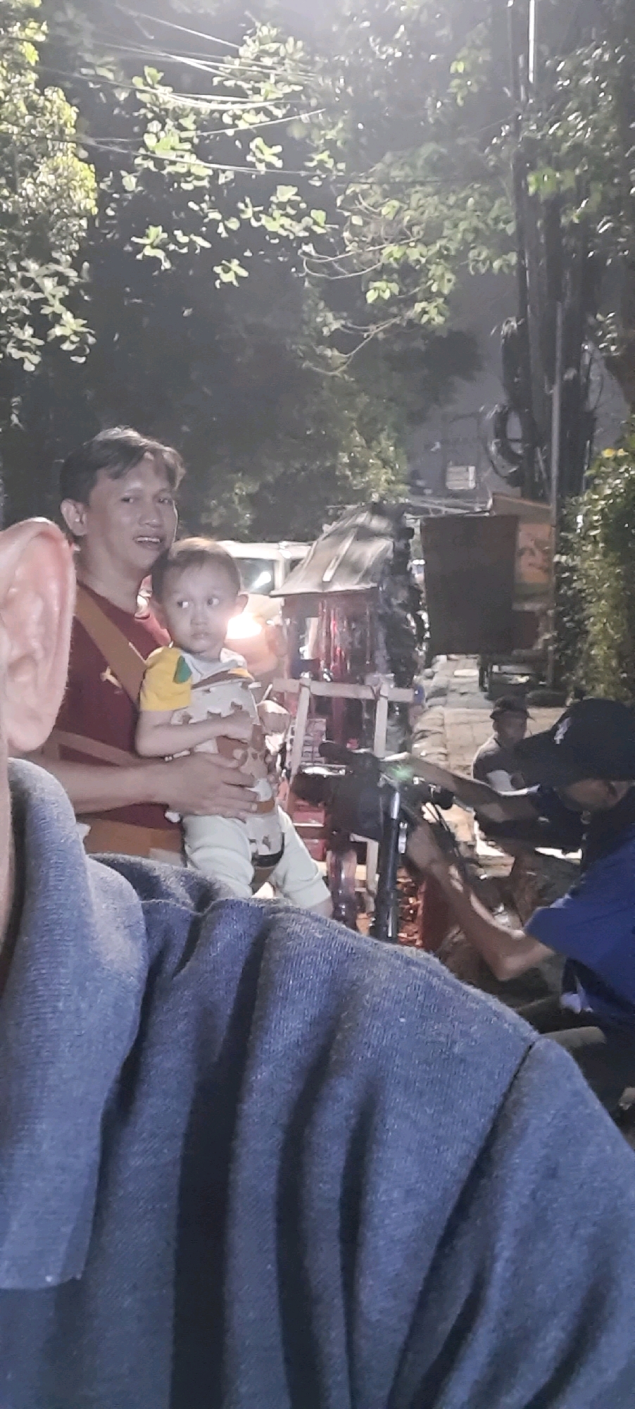 A low-light, slightly blurry medium shot shows a man holding a young child outdoors at night. The man, wearing a red shirt and a yellow strap across his chest, looks towards the left, his mouth slightly open. The child, in a yellow and patterned outfit, looks to the right with a neutral expression. In the foreground, the blue fabric of a garment, possibly a sweater, obscures the bottom left corner. The background features trees with illuminated leaves, a street scene with a vehicle, and several individuals. One person in a blue shirt and black cap is visible on the right, near a motorcycle. The scene appears to be on a street in East Jakarta, Indonesia, with the ambient lighting suggesting a public area at night.