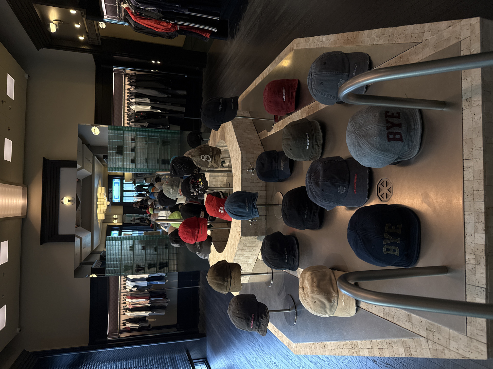 The image shows an interior view of a clothing store, with a display of various hats arranged on a table. The hats are of different colors and styles, some bearing the text "MASONPRINCE", "BYE", and "SPOKESHAR". The table has a textured surface, possibly stone or tile, and is partially obscured by metal bars. The hats are arranged on stands. Clothing racks line the walls of the store. The environment appears to be a retail setting, focusing on the display and sale of fashion items. The specific location within Liwan District, China cannot be ascertained from the image.
