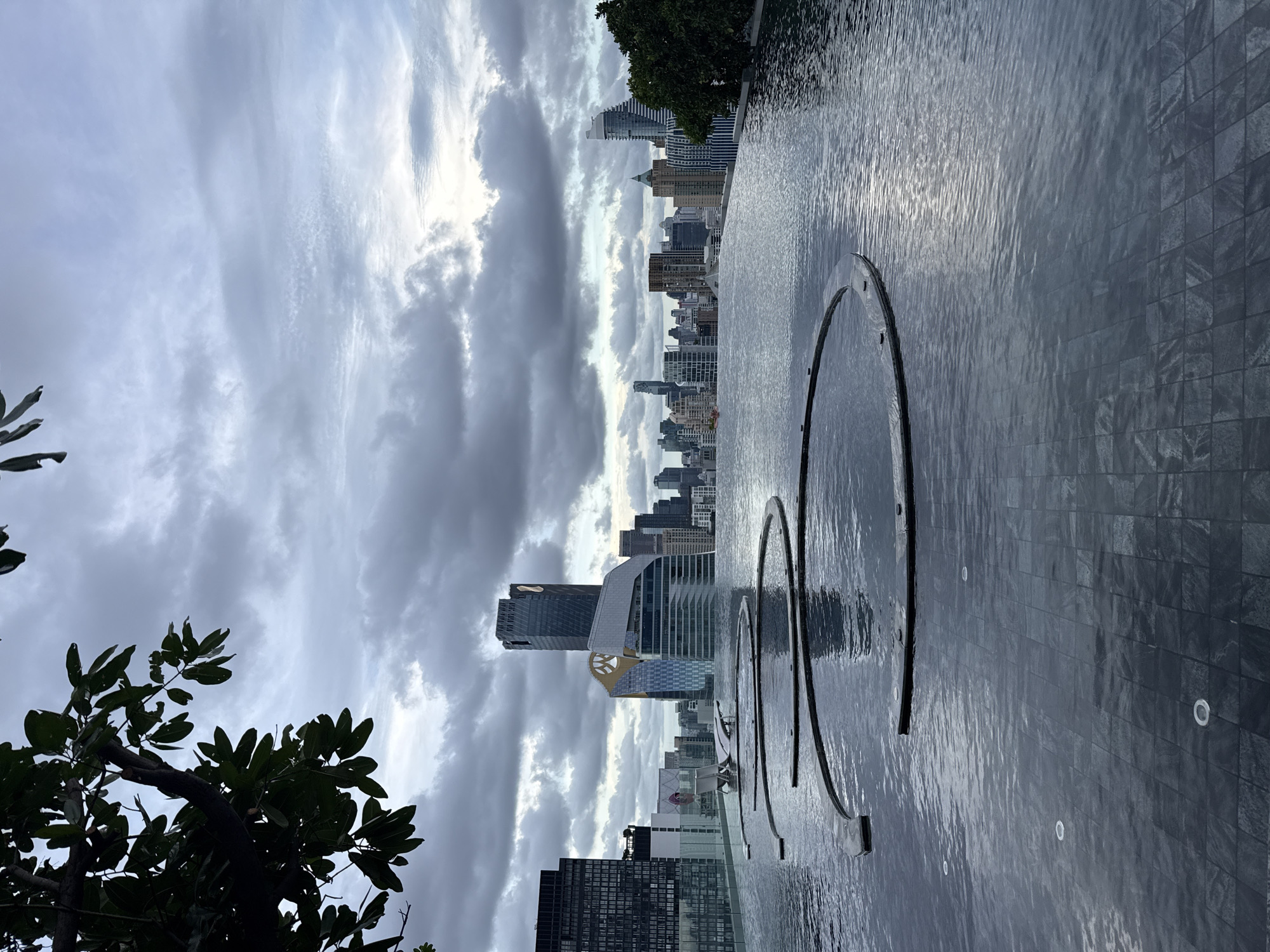 This is a vertical, eye-level, outdoor, medium shot of a rooftop infinity pool with a cityscape in the background. The pool's surface is rippled, reflecting the cloudy sky. Two circular, raised structures are visible in the water, one larger than the other, creating a contemporary design element. The pool's edge is lined with dark, tiled material.
In the background, a dense cityscape of high-rise buildings stretches across the horizon. The buildings vary in height and architectural style, with some featuring glass facades and distinctive shapes. A large tree with dark green leaves is silhouetted in the lower left corner, partially obscuring the view of the cityscape.
The sky is overcast with dramatic, heavy clouds, suggesting impending rain or recent precipitation. The lighting is diffused, indicating either early morning or late afternoon. No people are visible in the image. The overall mood is serene yet imposing, with the vastness of the city juxtaposed against the tranquility of the pool. The location cues point to Bangkok, Thailand, given the dense urban environment and architectural styles common in the city.